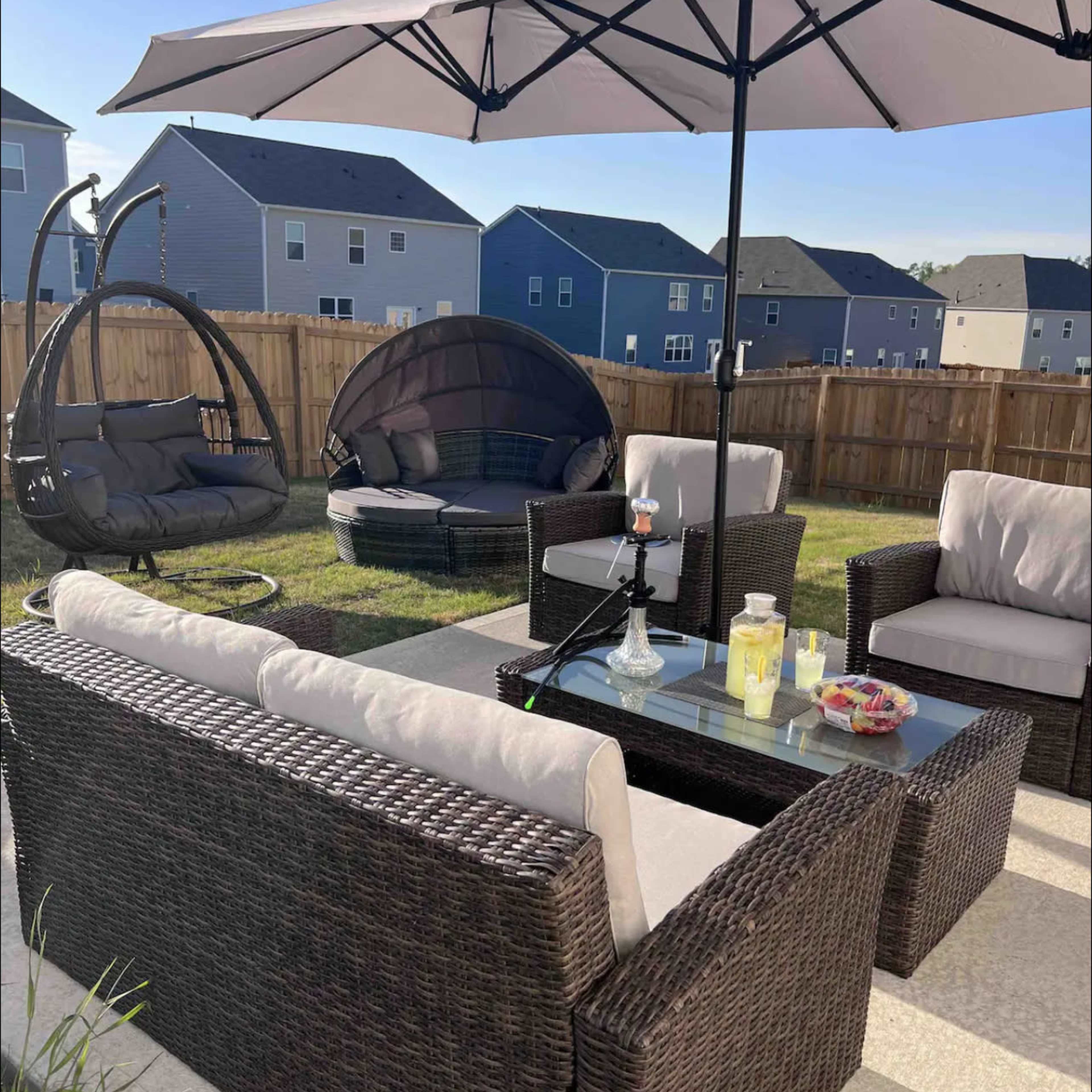 An outdoor patio area with a seating arrangement that includes wicker furniture, a glass coffee table, and a large umbrella, set against a wooden fence and several residential homes in the background.