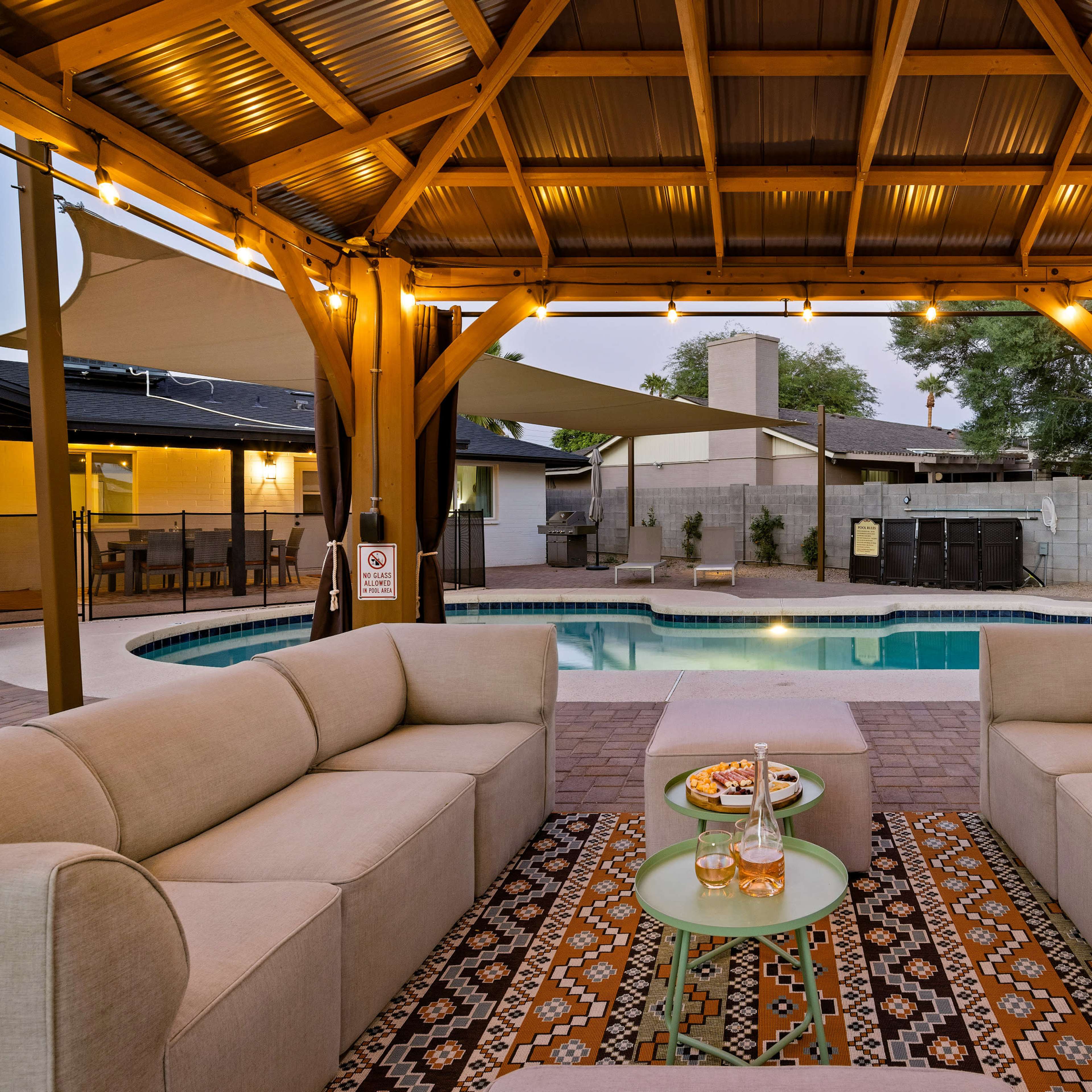 A covered outdoor seating area features light-colored sofas and a small table beside a pool surrounded by a fenced yard.