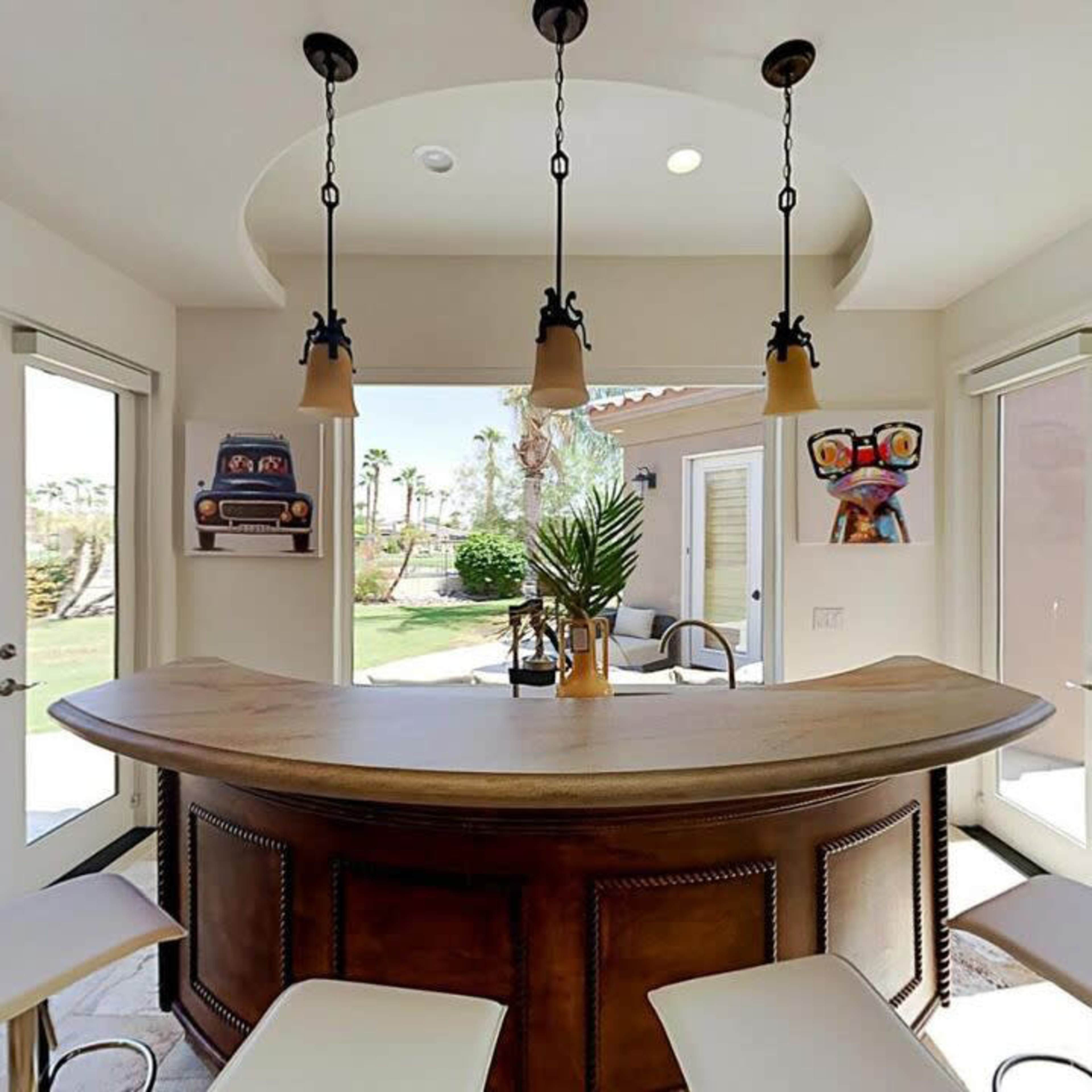 A curved wooden bar with pendant lights is situated in a bright room surrounded by glass doors leading to a garden.