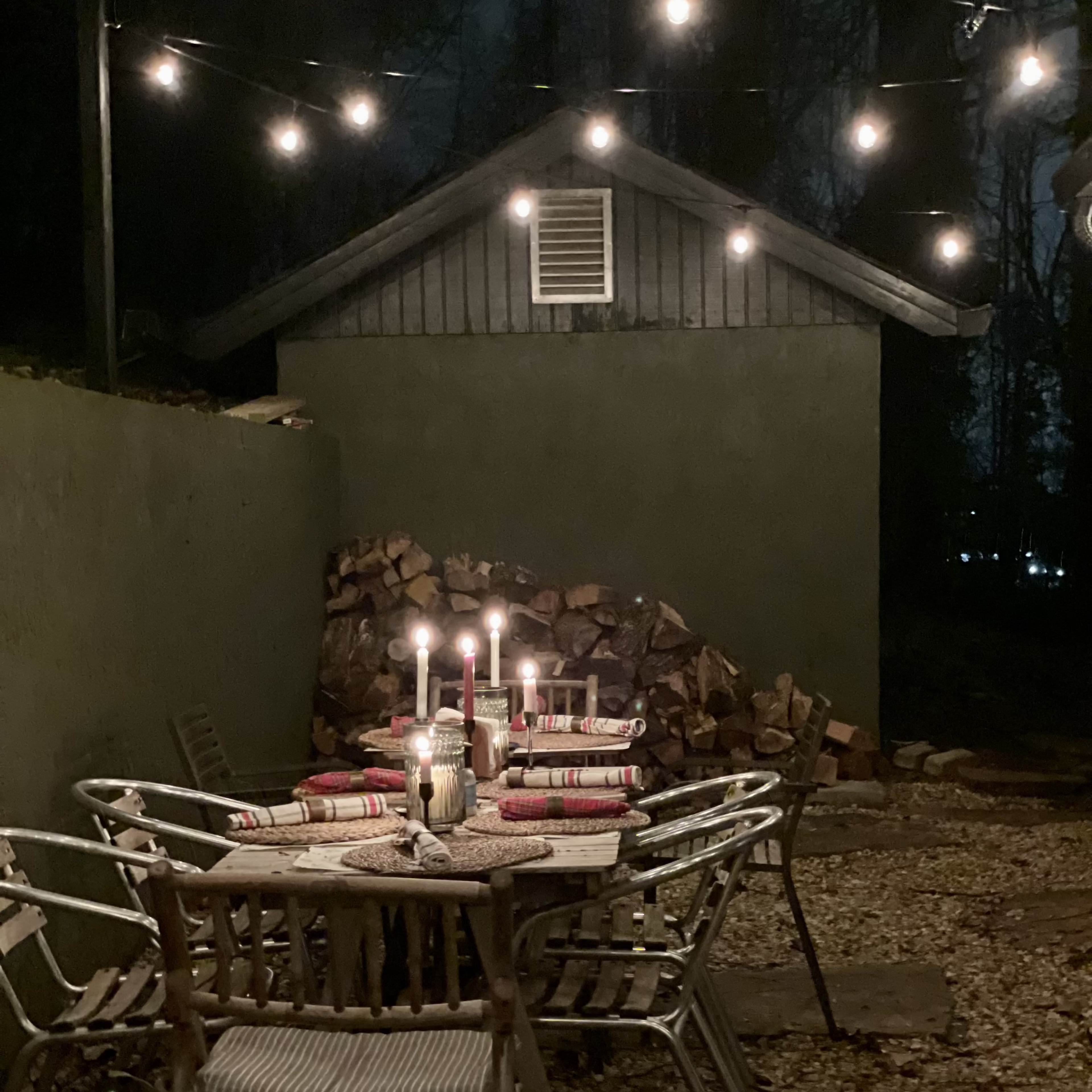 A dimly lit outdoor dining area features a table set with candles and surrounded by chairs, with a stack of firewood and string lights in the background.