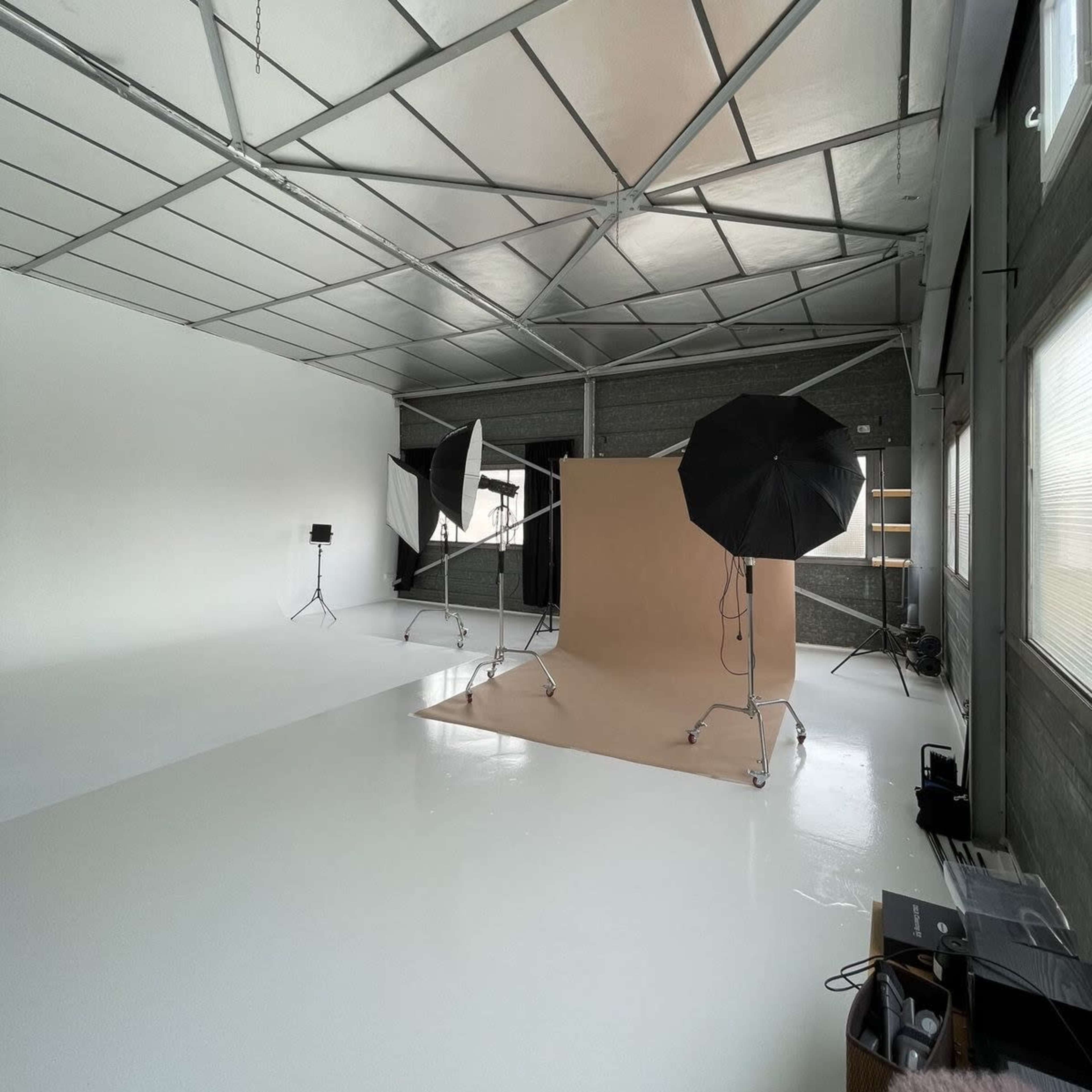 A spacious photography studio with a beige backdrop, three lighting setups, and white polished floors, all under a metal ceiling.