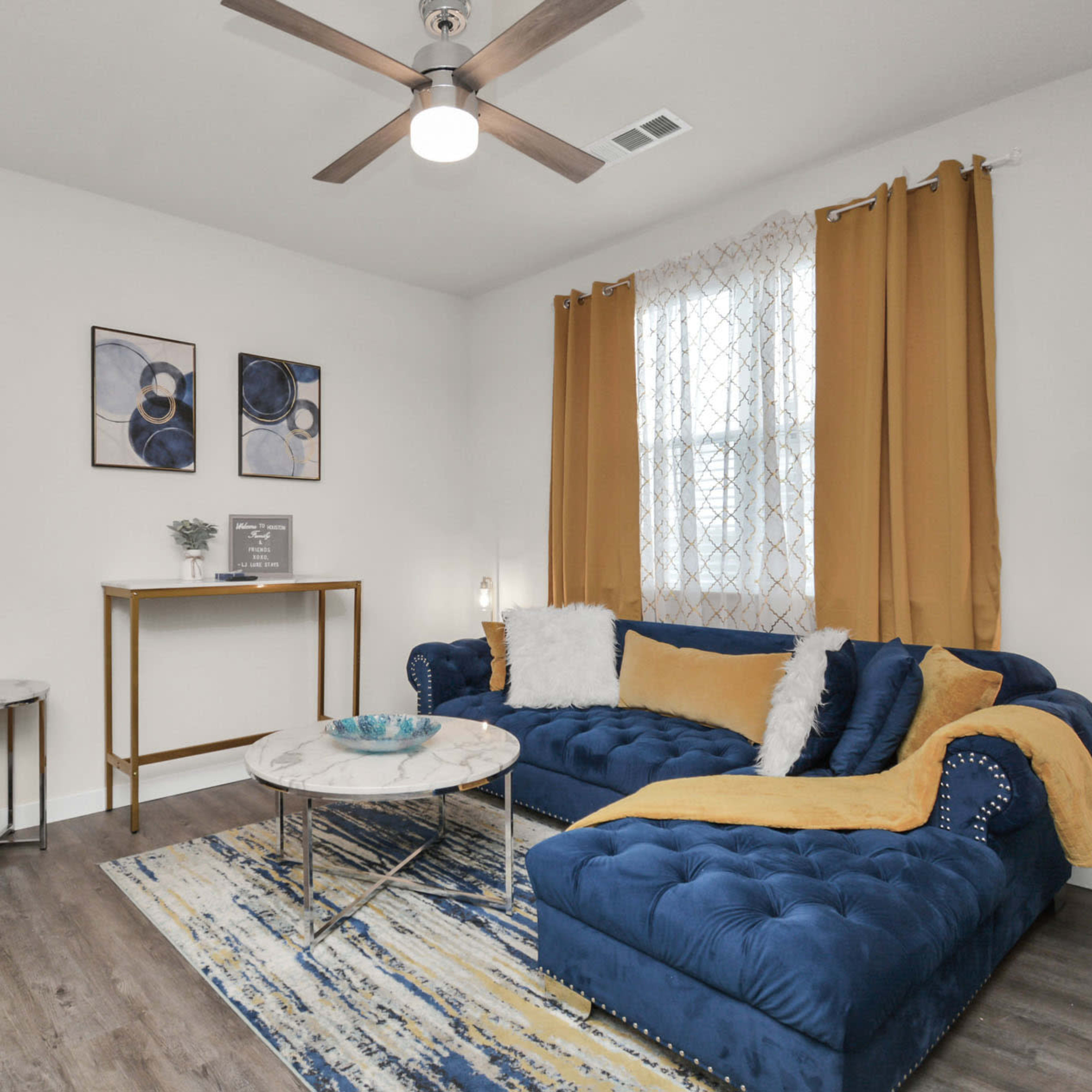 The living room features a blue sectional sofa, a round marble coffee table, a yellow armchair, and large yellow curtains.