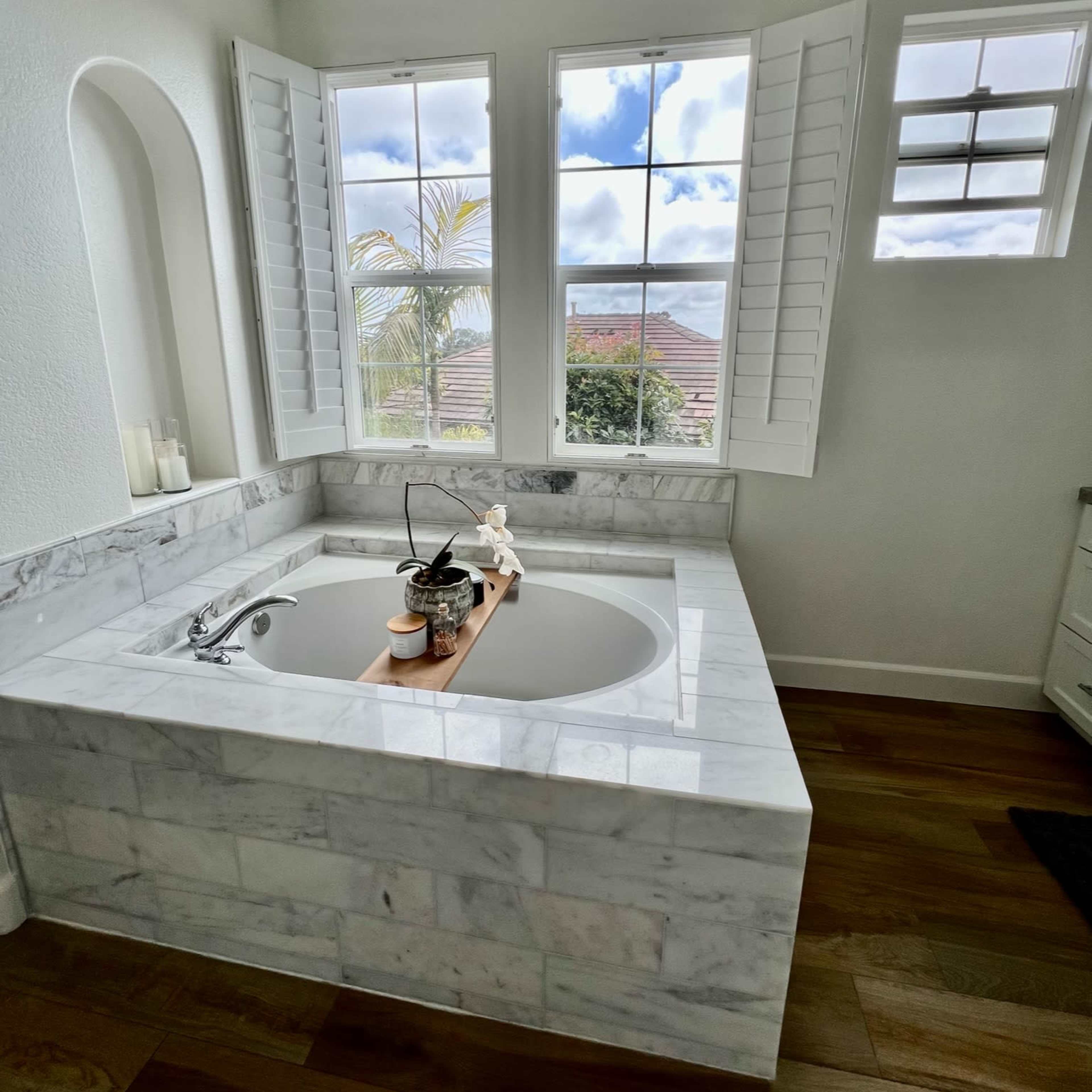 A well-lit bathroom featuring a marble bathtub with a wooden tray in front of large windows with plantation shutters.