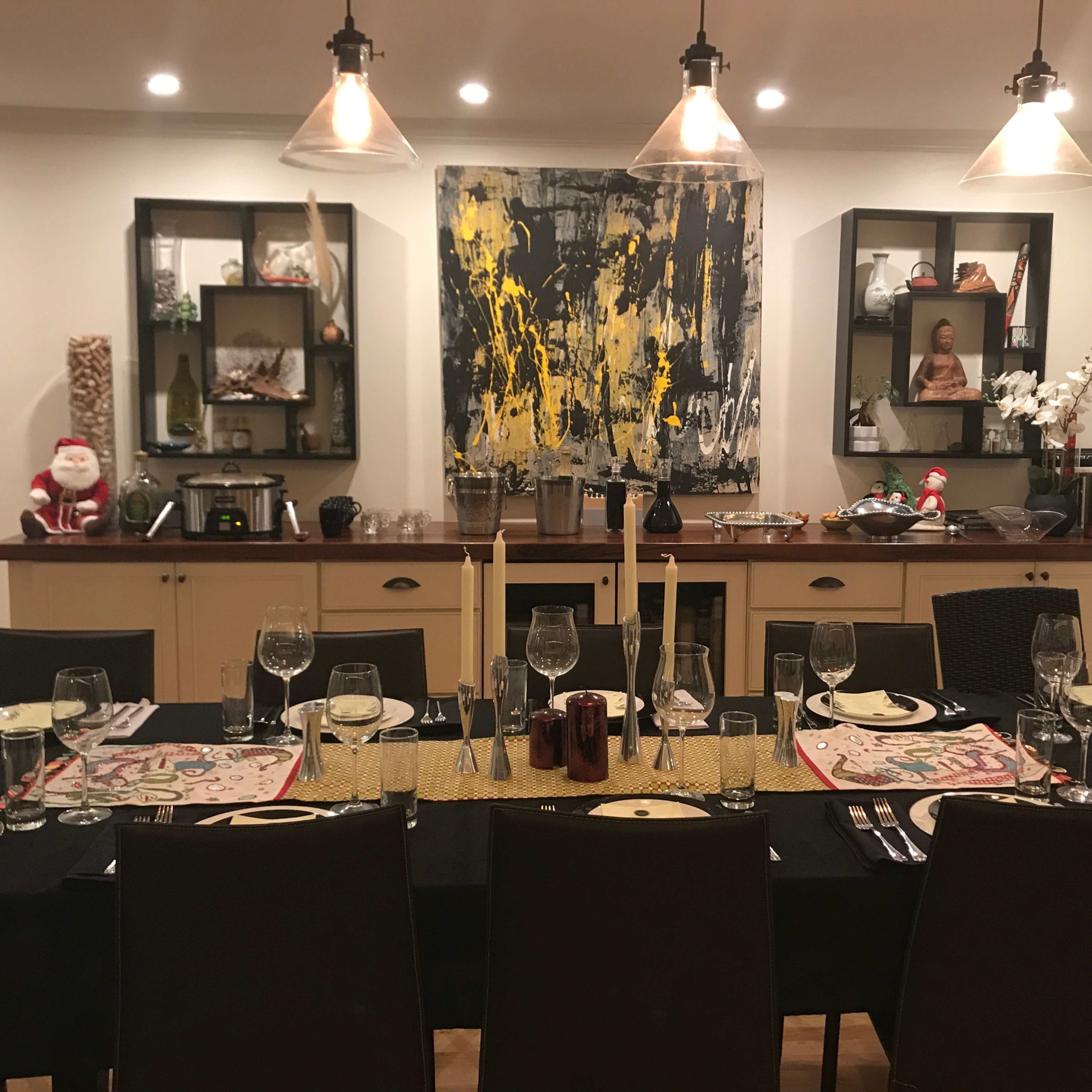A dining table is set with glassware and placemats, positioned in a brightly lit room featuring a large artwork and shelves with decorative items.