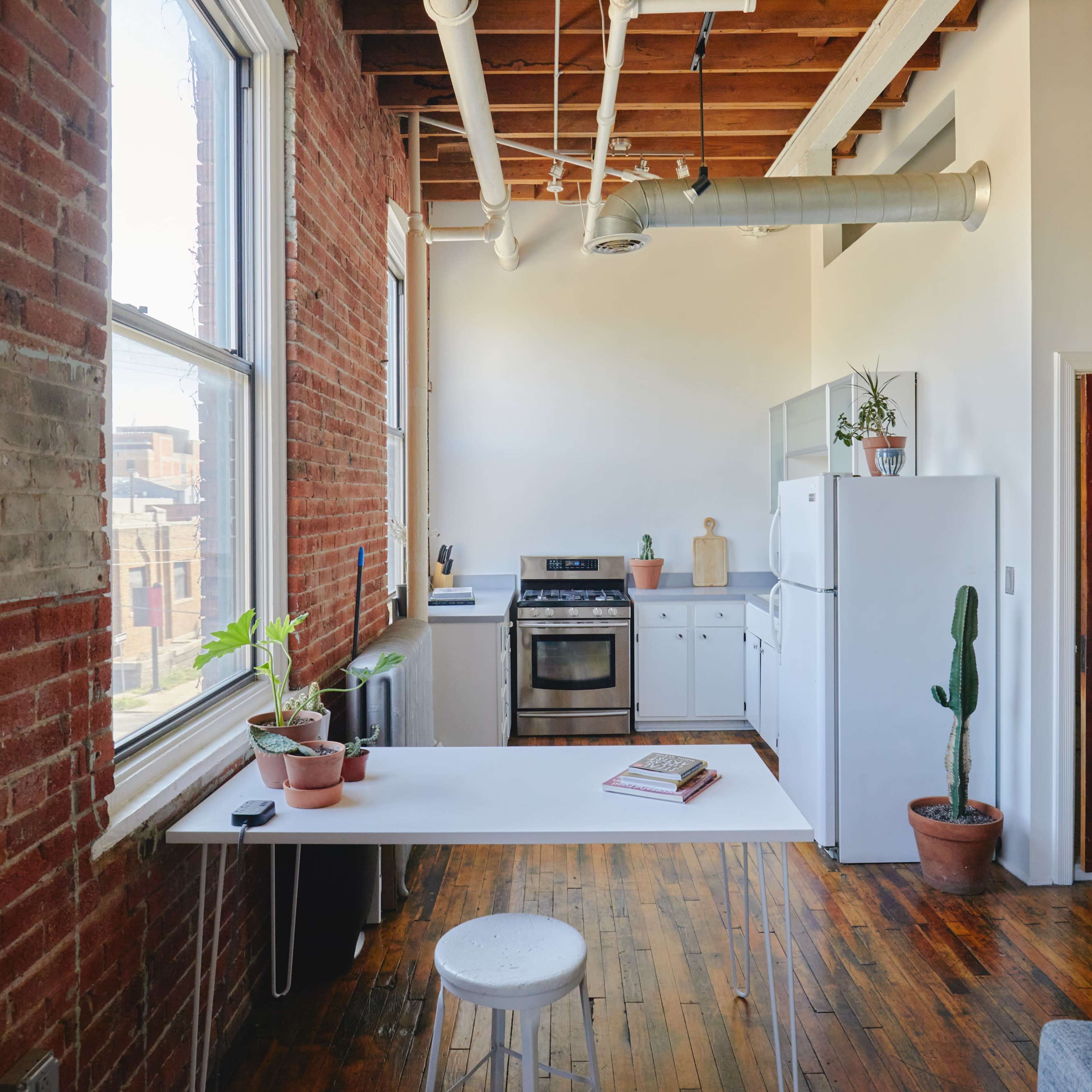 A bright kitchen area features a white table, a stove, a refrigerator, and potted plants against exposed brick walls and wooden flooring.