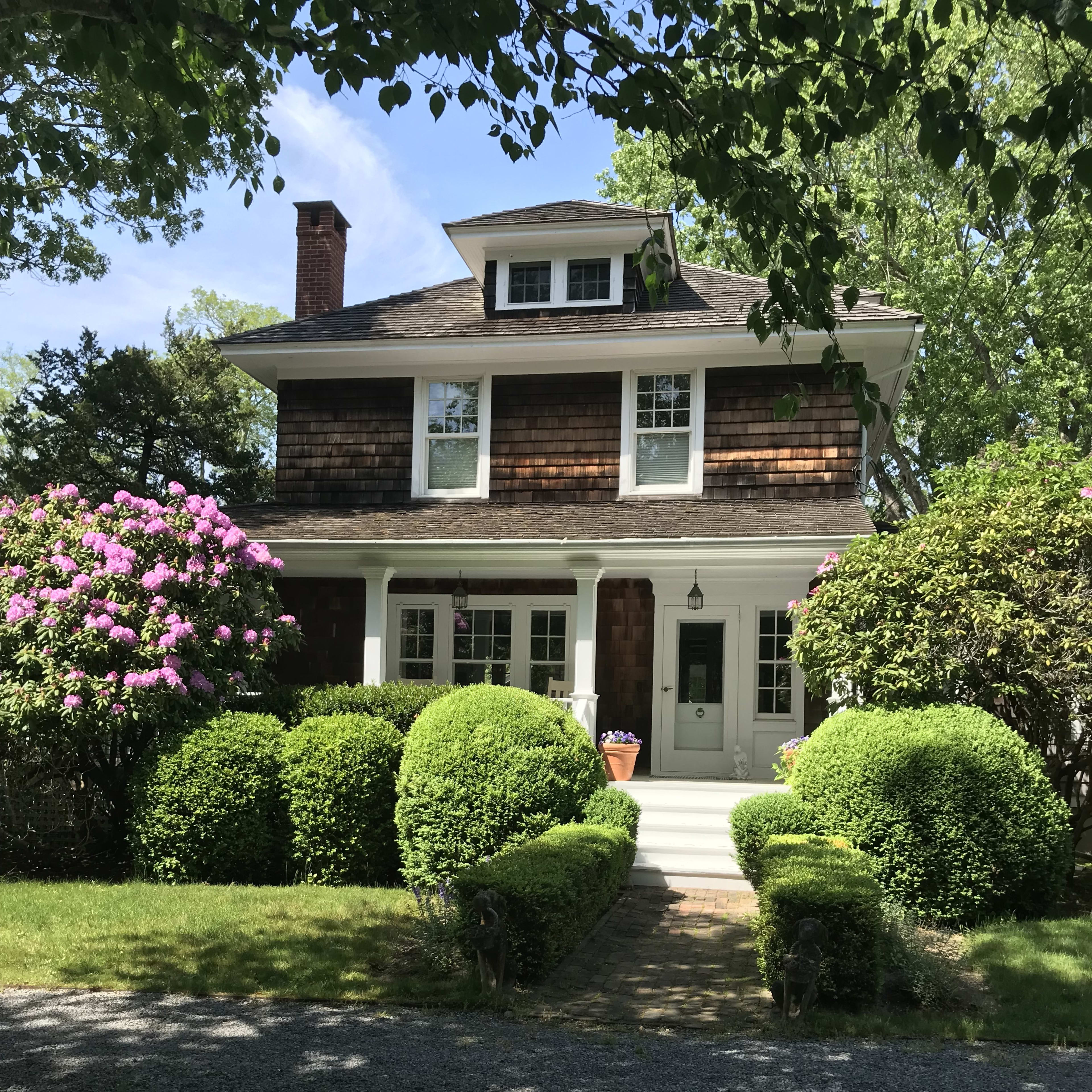 A two-story shingle-style house with a front porch, surrounded by neatly trimmed bushes and flowering plants, set on a gravel driveway.