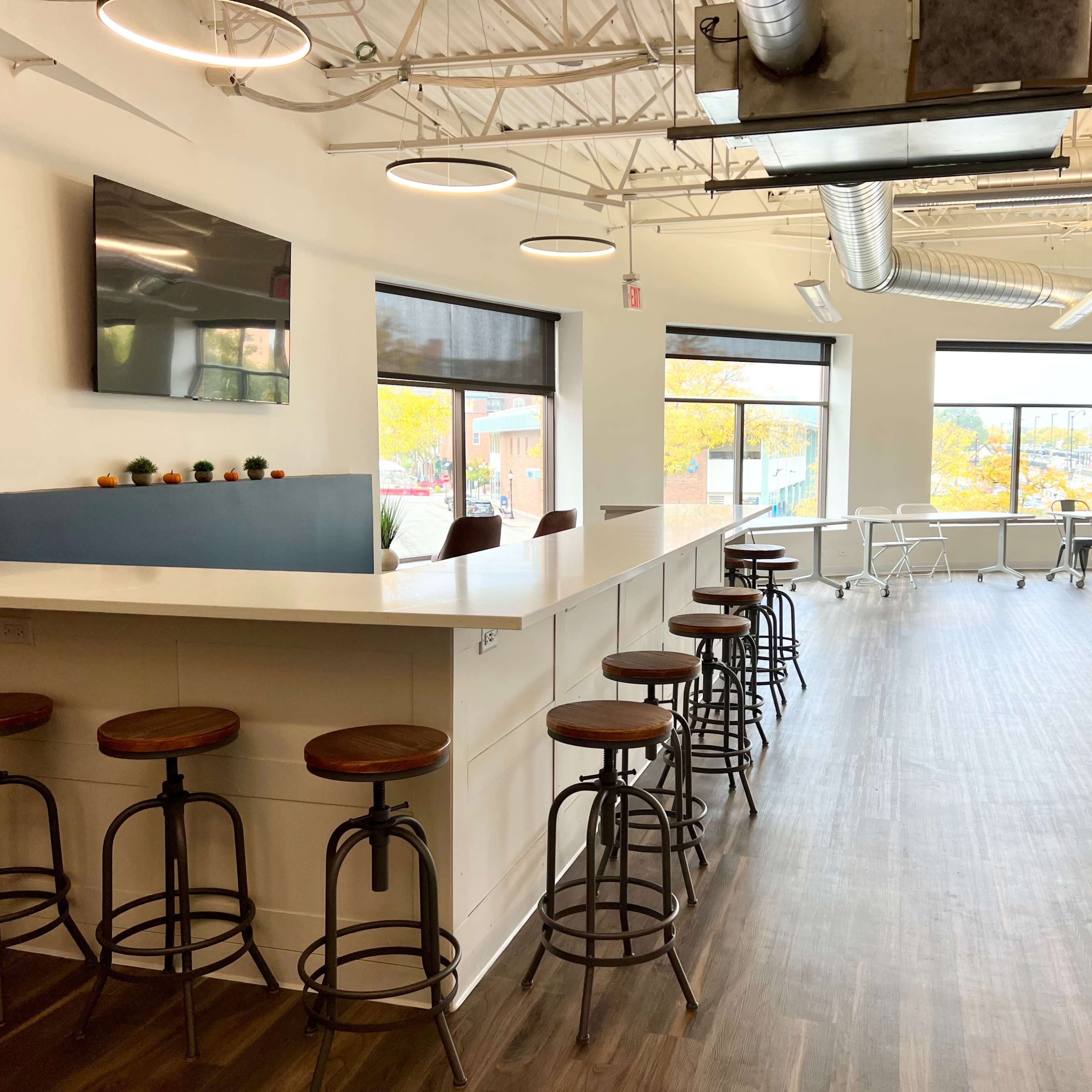 A modern office interior with a bar-height counter and several stools on one side, complemented by large windows letting in natural light.