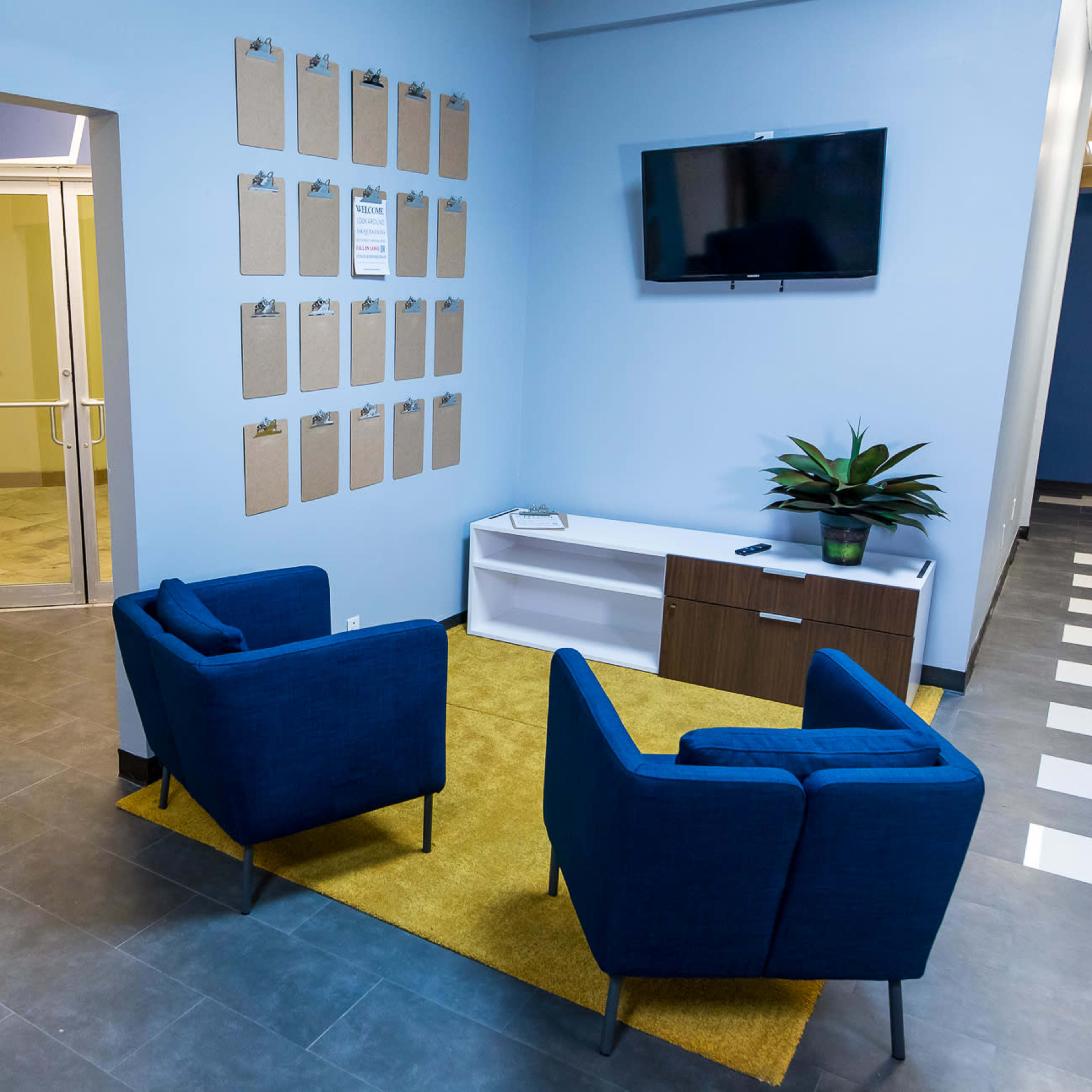 A modern waiting area with two blue armchairs, a white cabinet, a wall-mounted television, and a number of clipboards arranged on the wall.