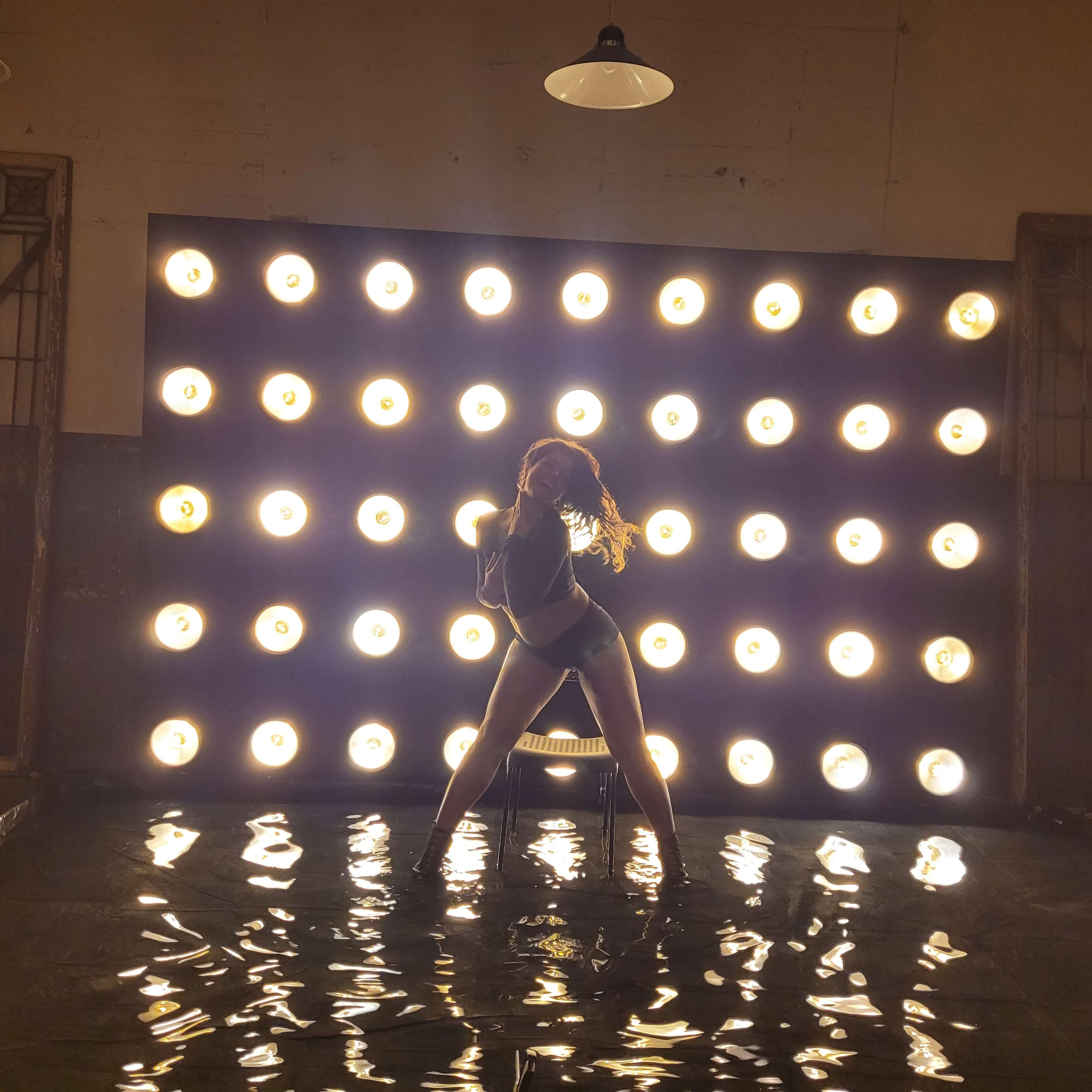 A dancer poses in front of a wall of bright lights, with water pooling at their feet.