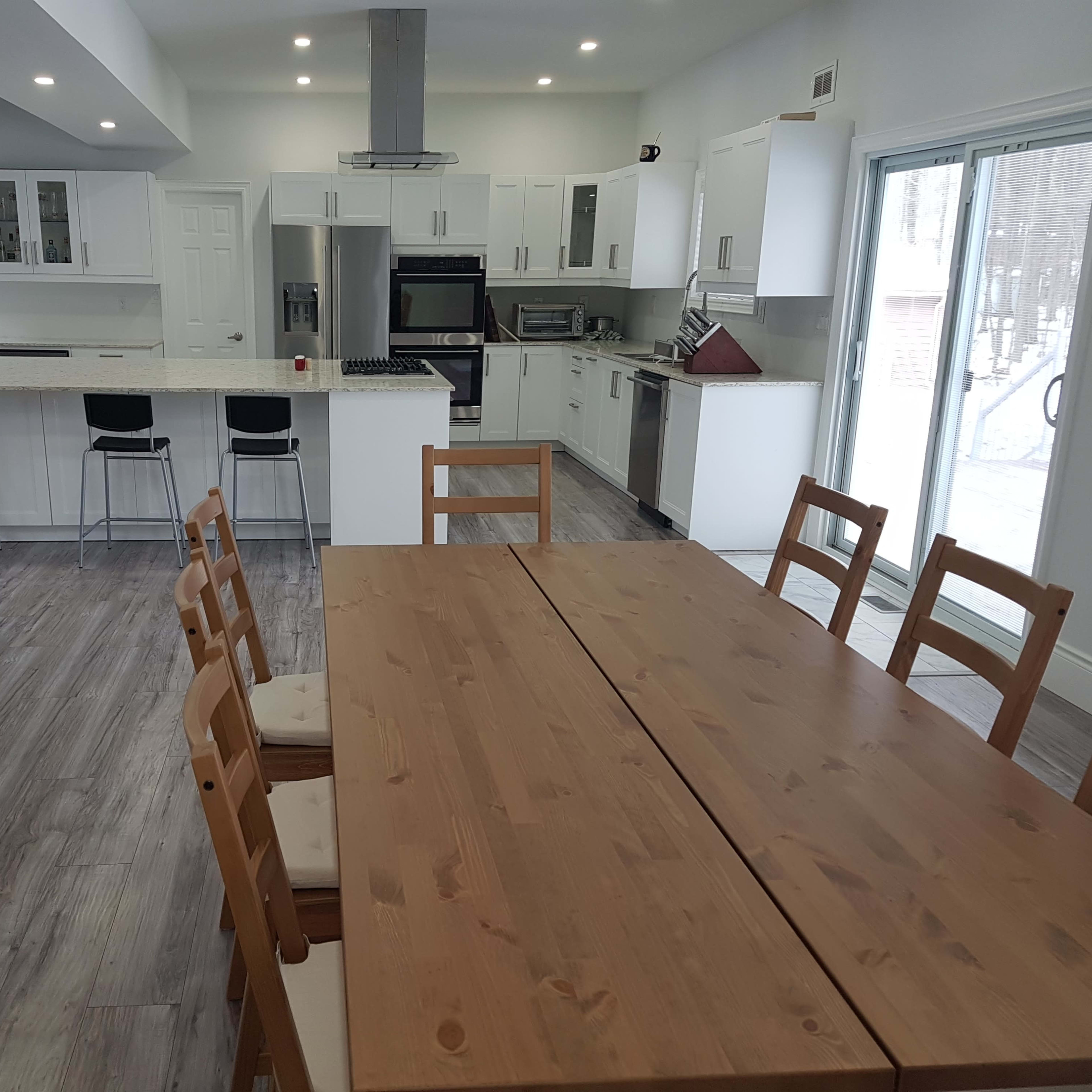 A spacious kitchen and dining area feature a large wooden table surrounded by chairs, with modern appliances and white cabinetry visible in the background.