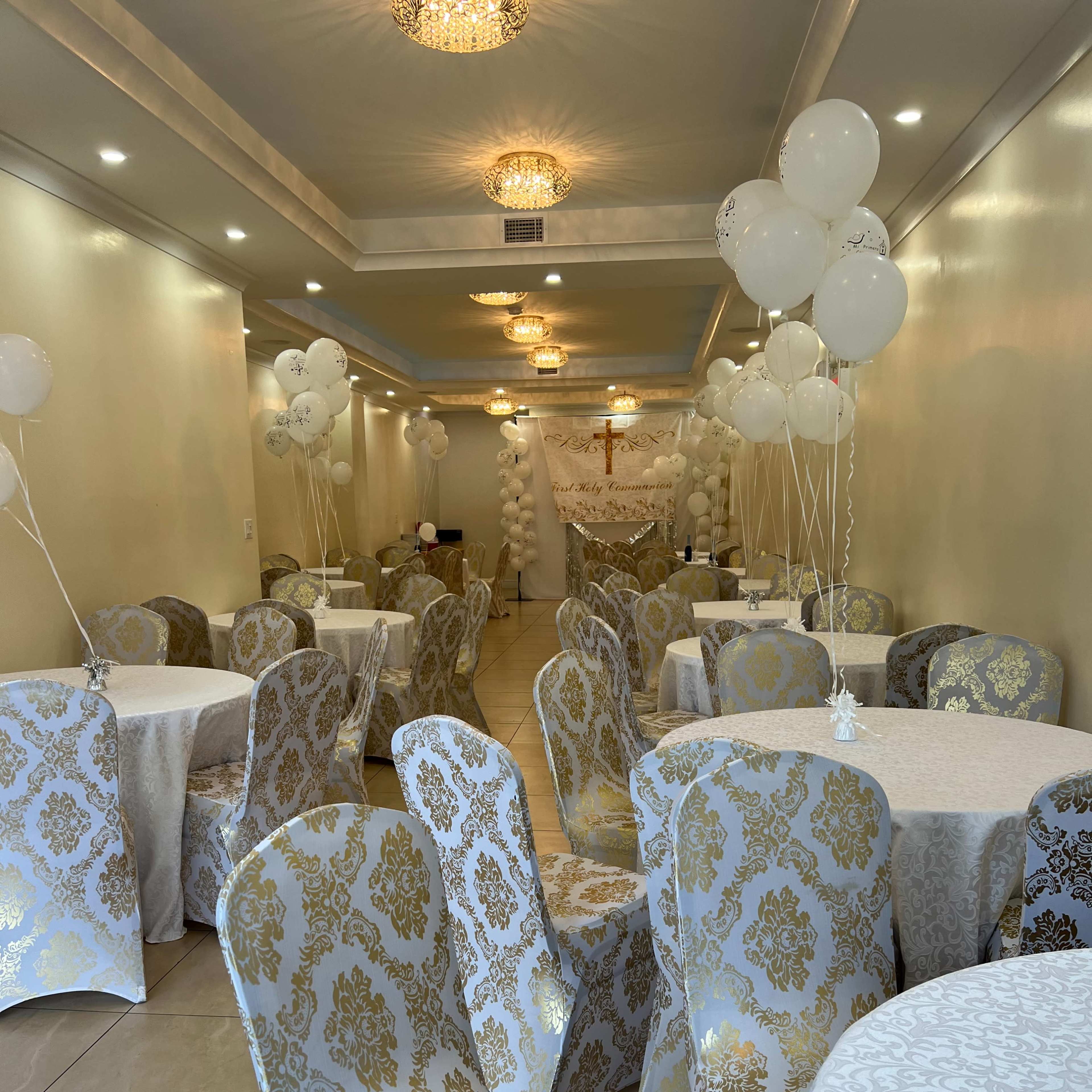 A decorated event space with tables covered in white and gold tablecloths, adorned with matching chairs and white balloons.