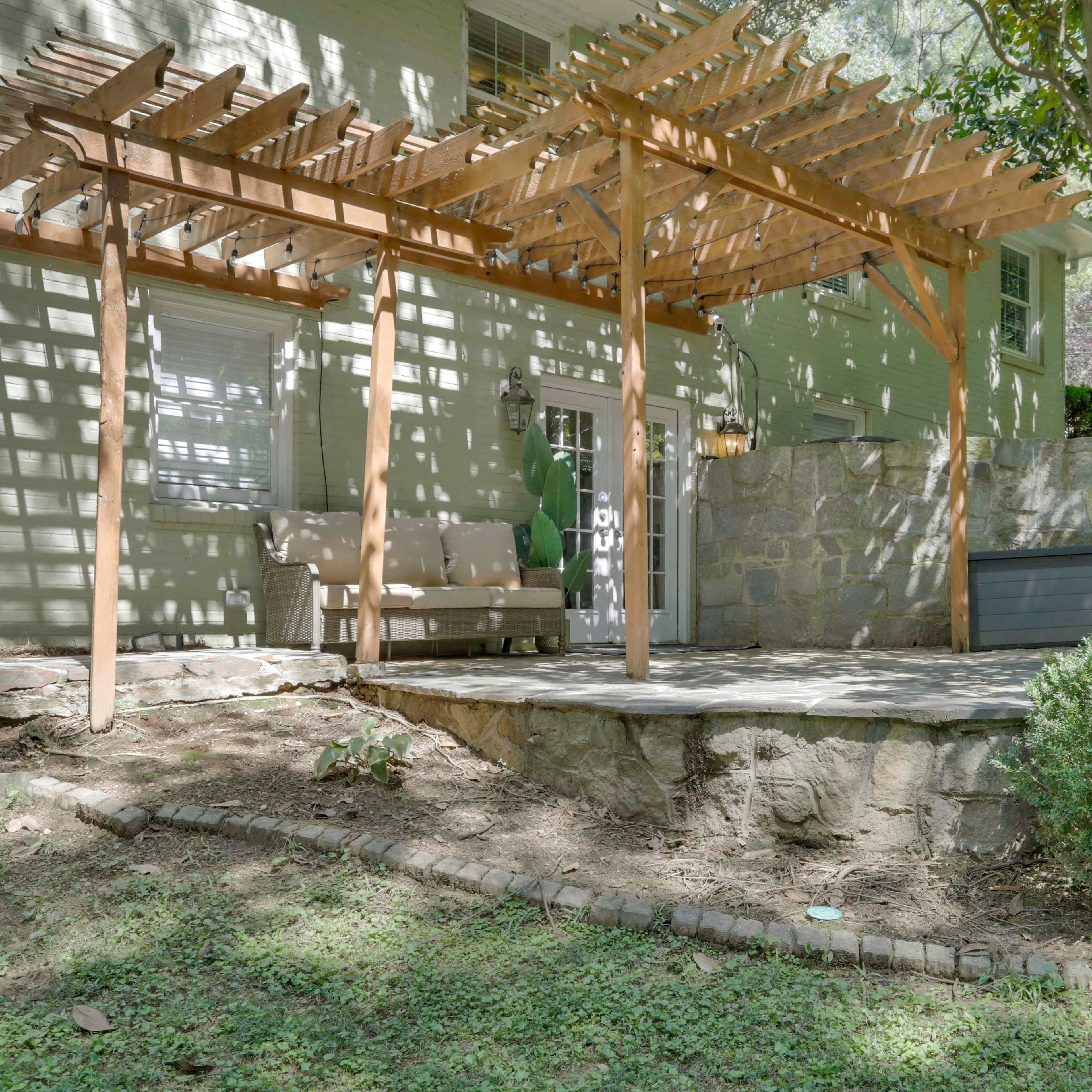 A patio area with a wooden pergola, a stone wall, and neatly trimmed shrubs in a residential backyard.