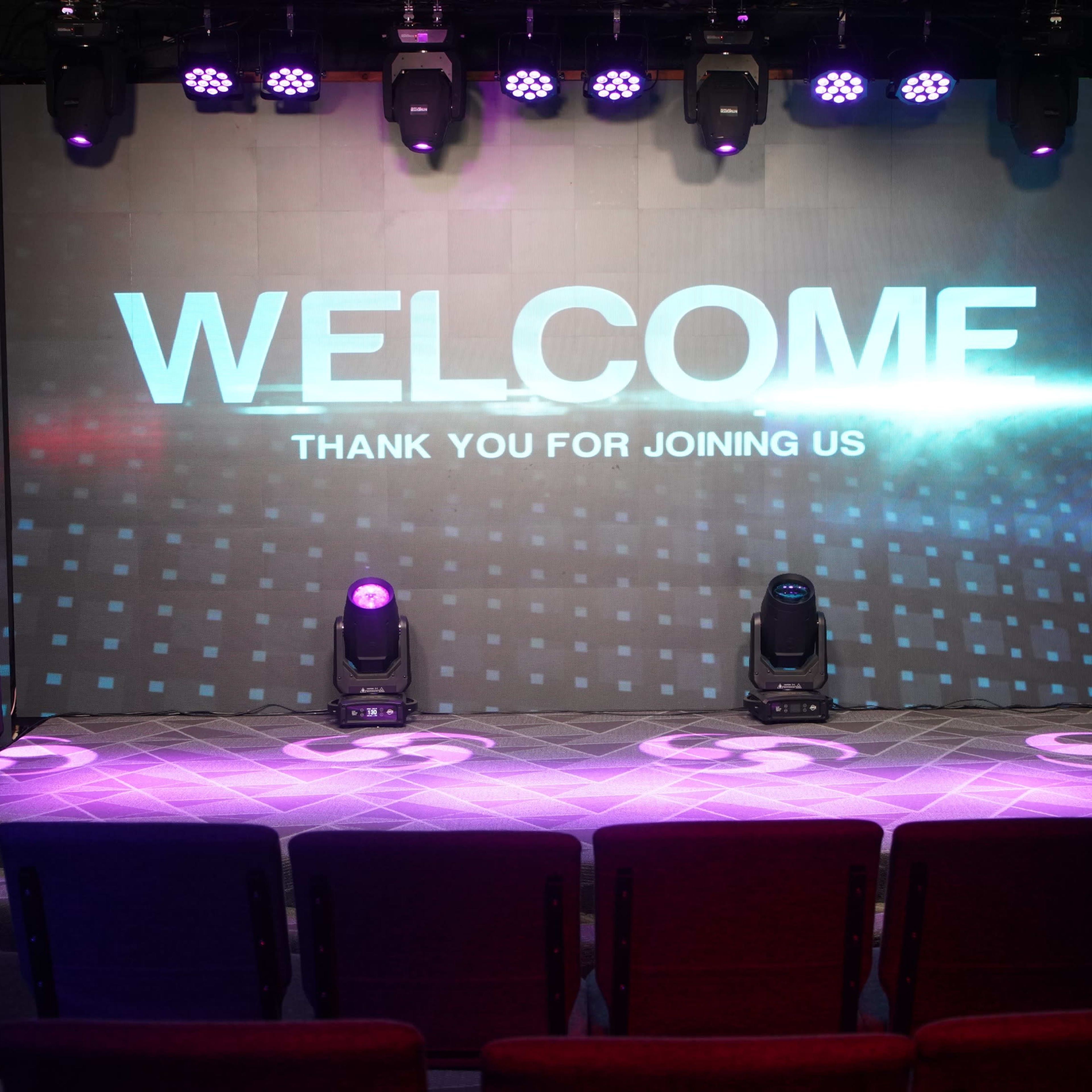 A stage is set with a large screen displaying the words "WELCOME" and "THANK YOU FOR JOINING US," surrounded by purple and white lighting.