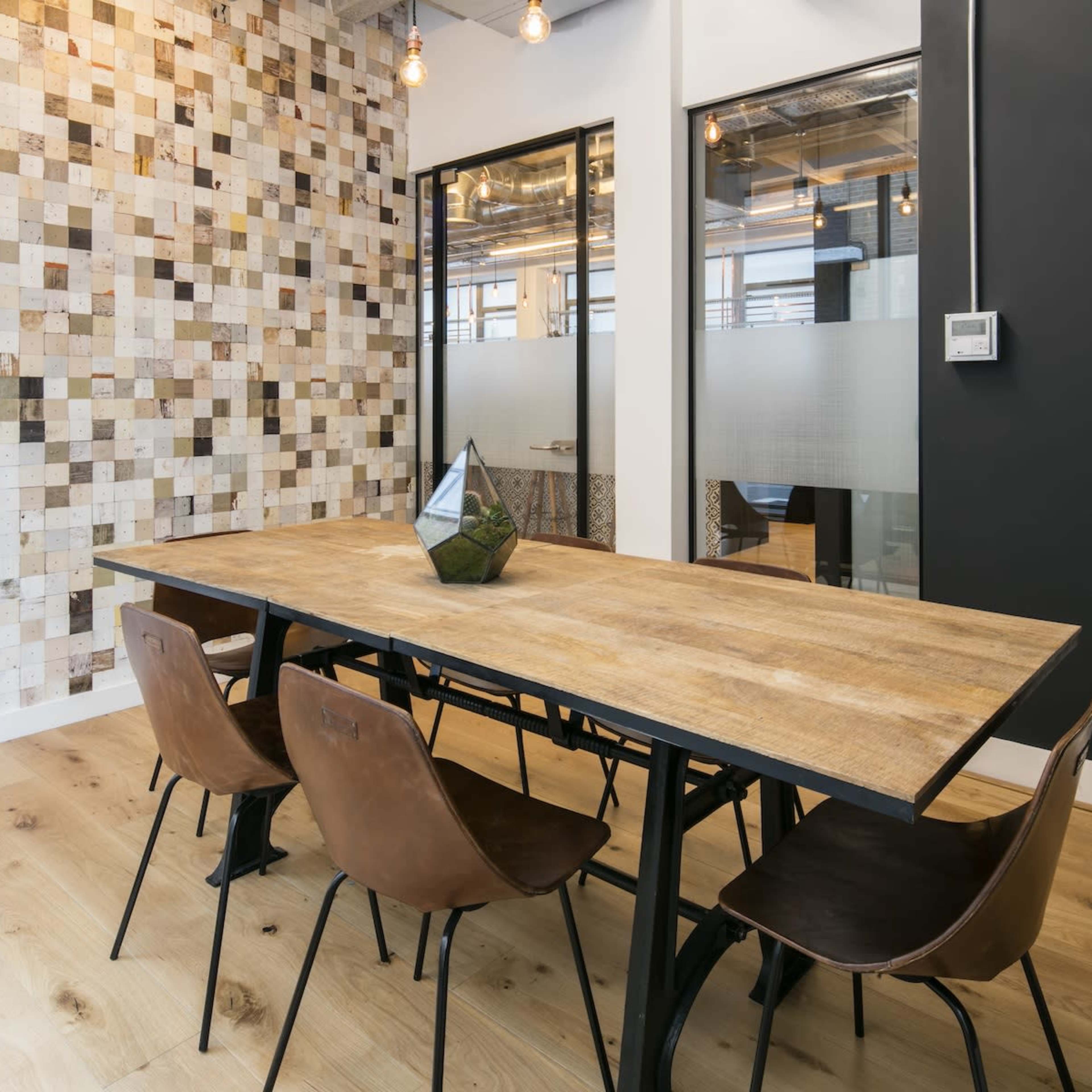 A wooden table with metal legs is set for dining in a room featuring a mosaic wall and glass-paneled office entrances.