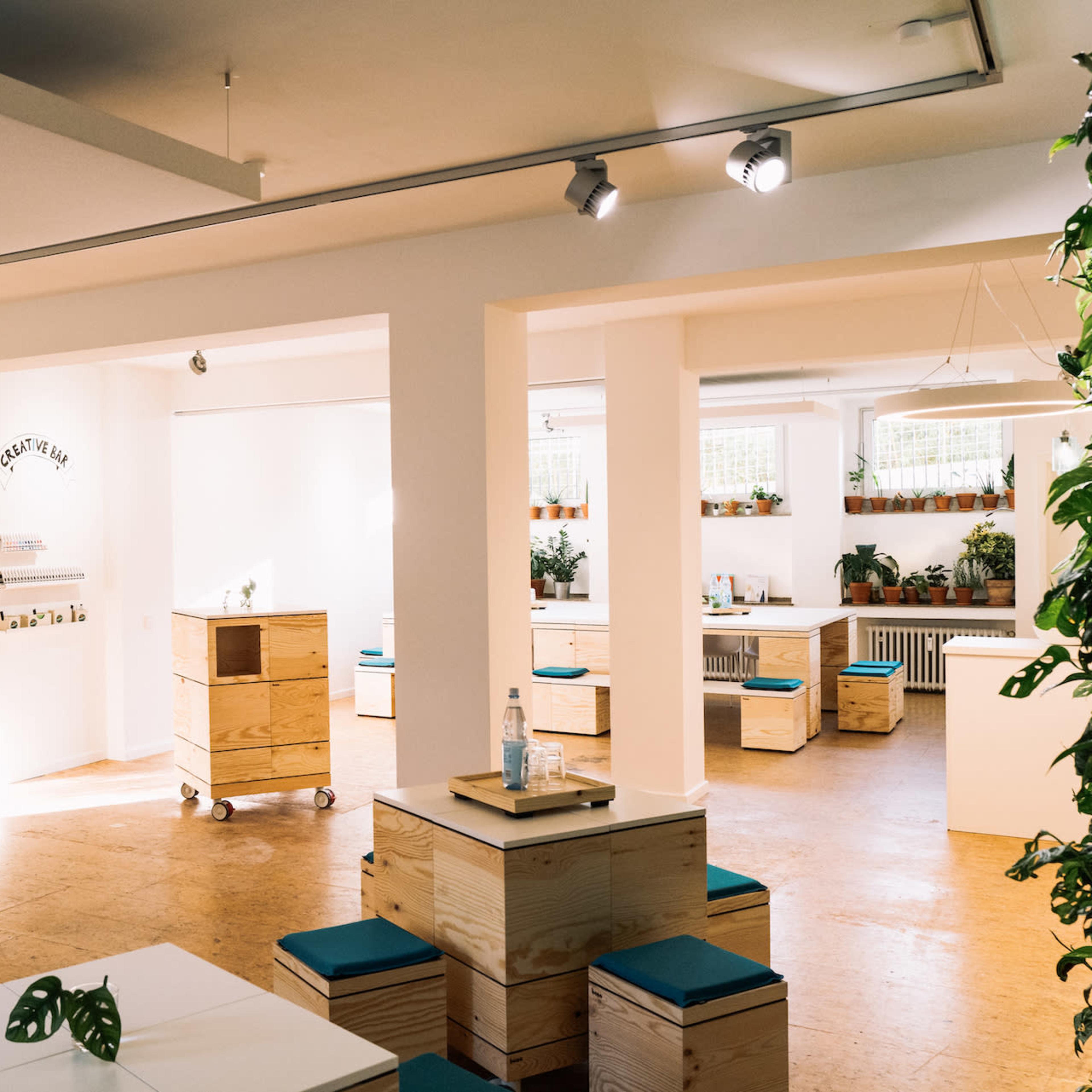 A bright, open workspace featuring wooden boxes and tables arranged around potted plants and walls adorned with colorful notes.