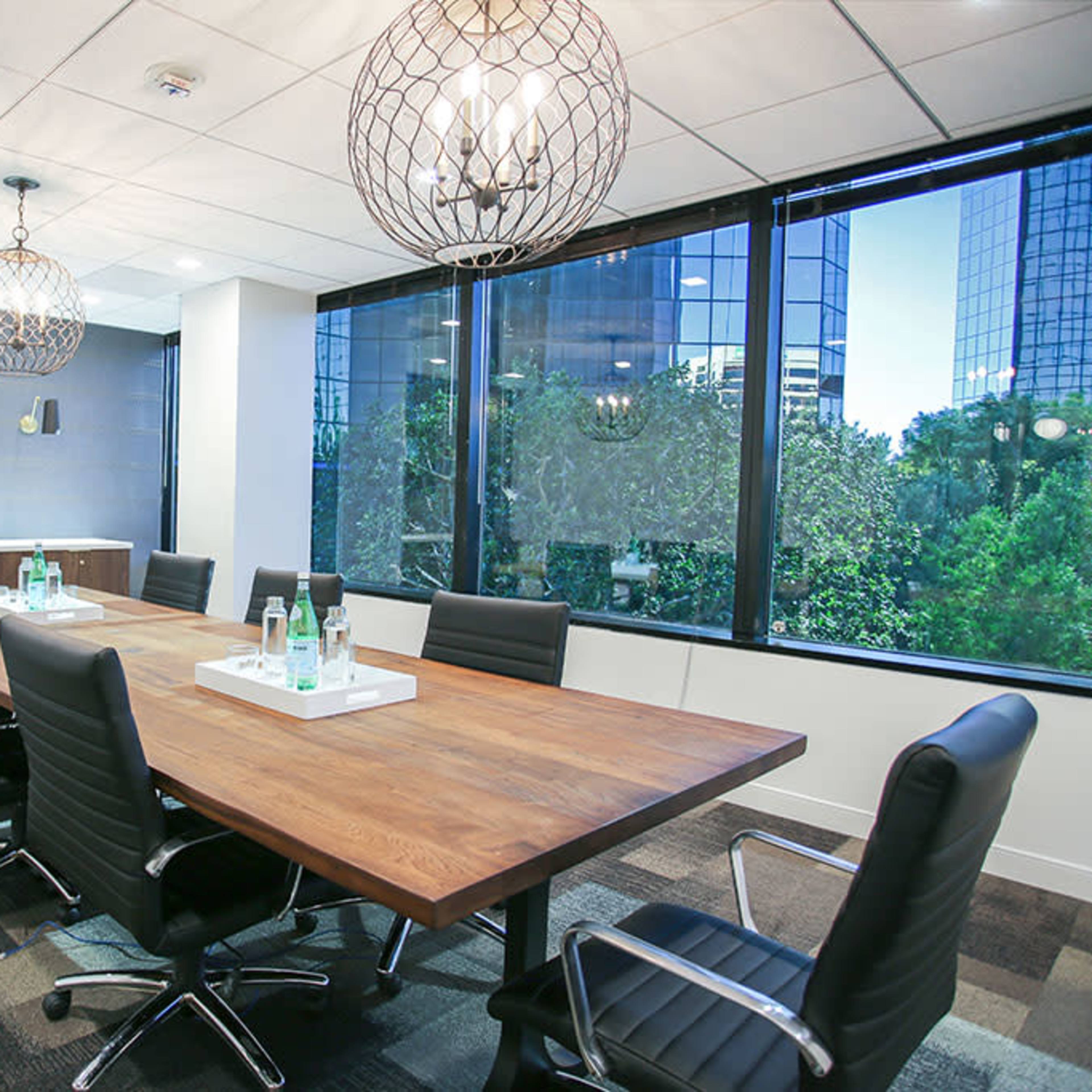 A modern conference room features a long wooden table surrounded by black chairs, with large windows providing a view of greenery outside.