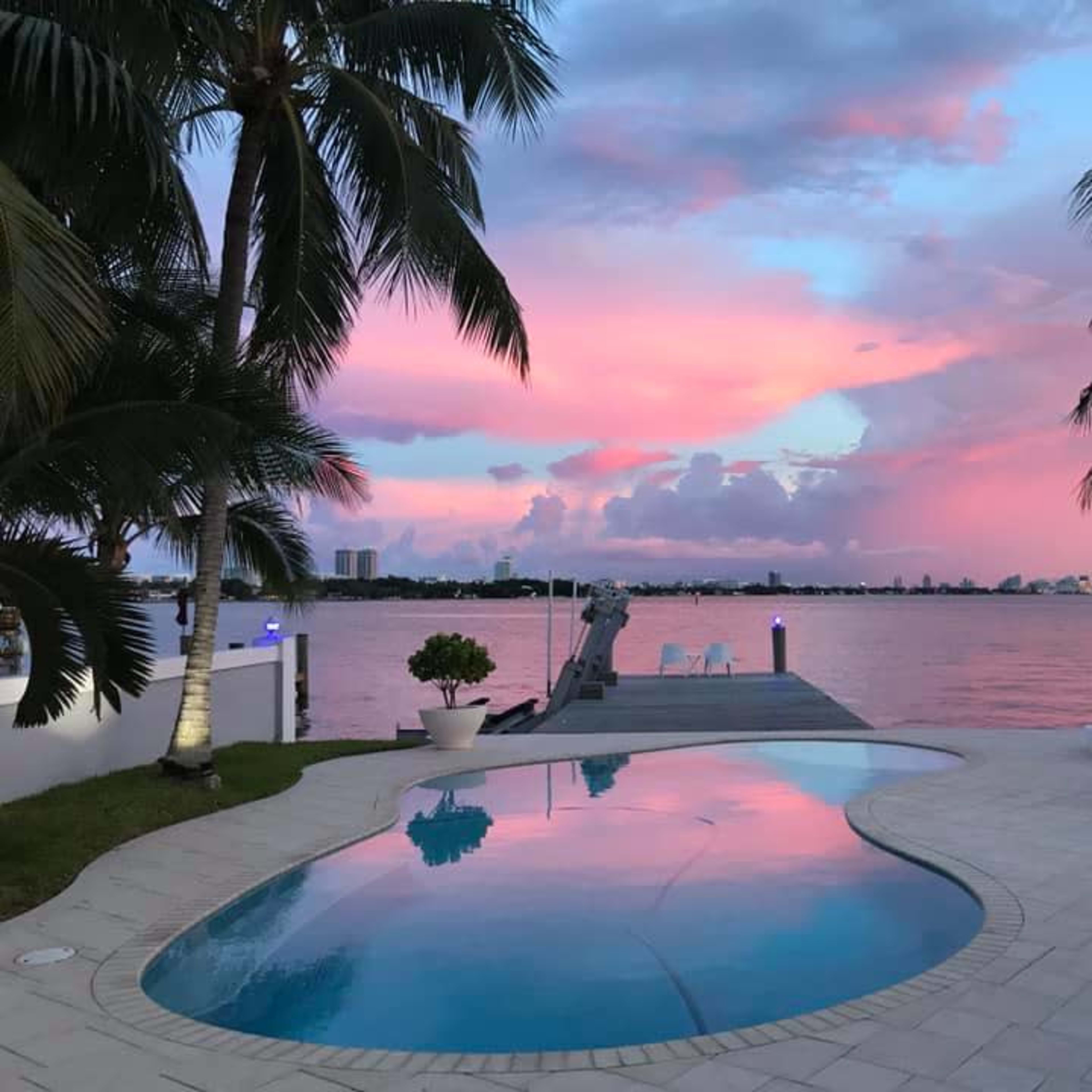 The scene shows a poolside area with palm trees and a waterfront view, featuring a vibrant pink and purple sky reflecting in the water.