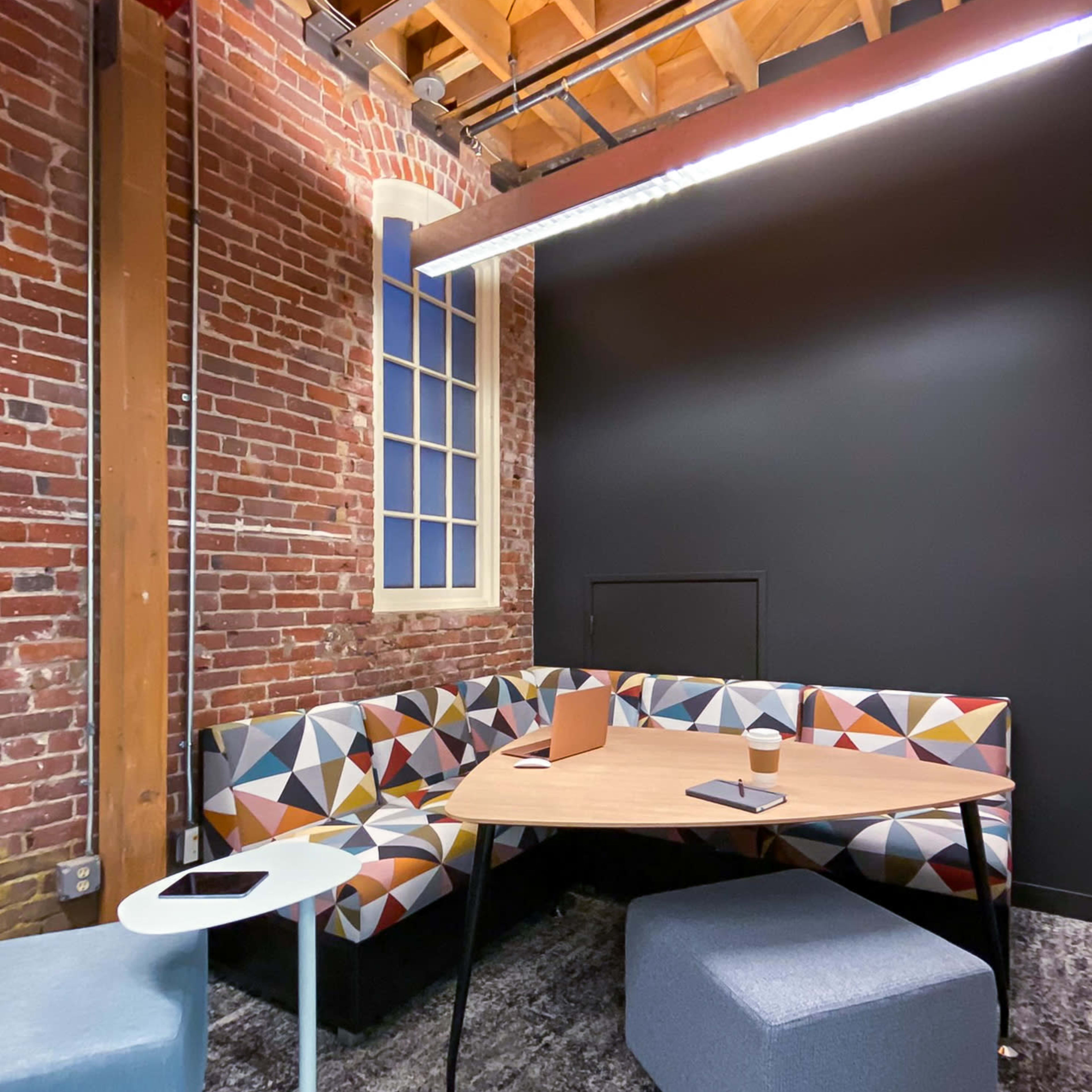 A modern workspace featuring a patterned seating area, a wooden table, and small stools, with exposed brick walls and a dark backdrop.