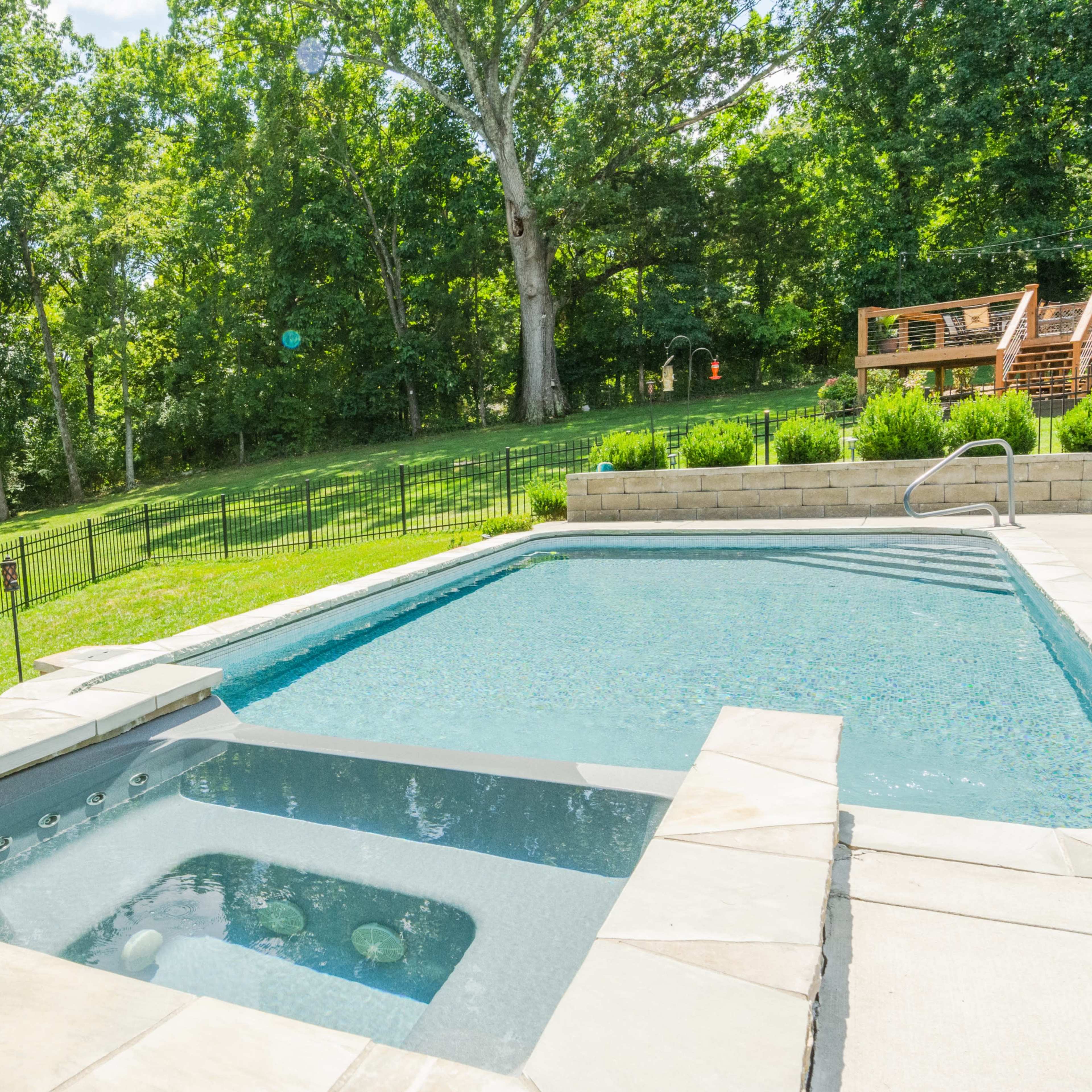 A backyard with a swimming pool and a spa area, surrounded by a fenced lawn and trees.