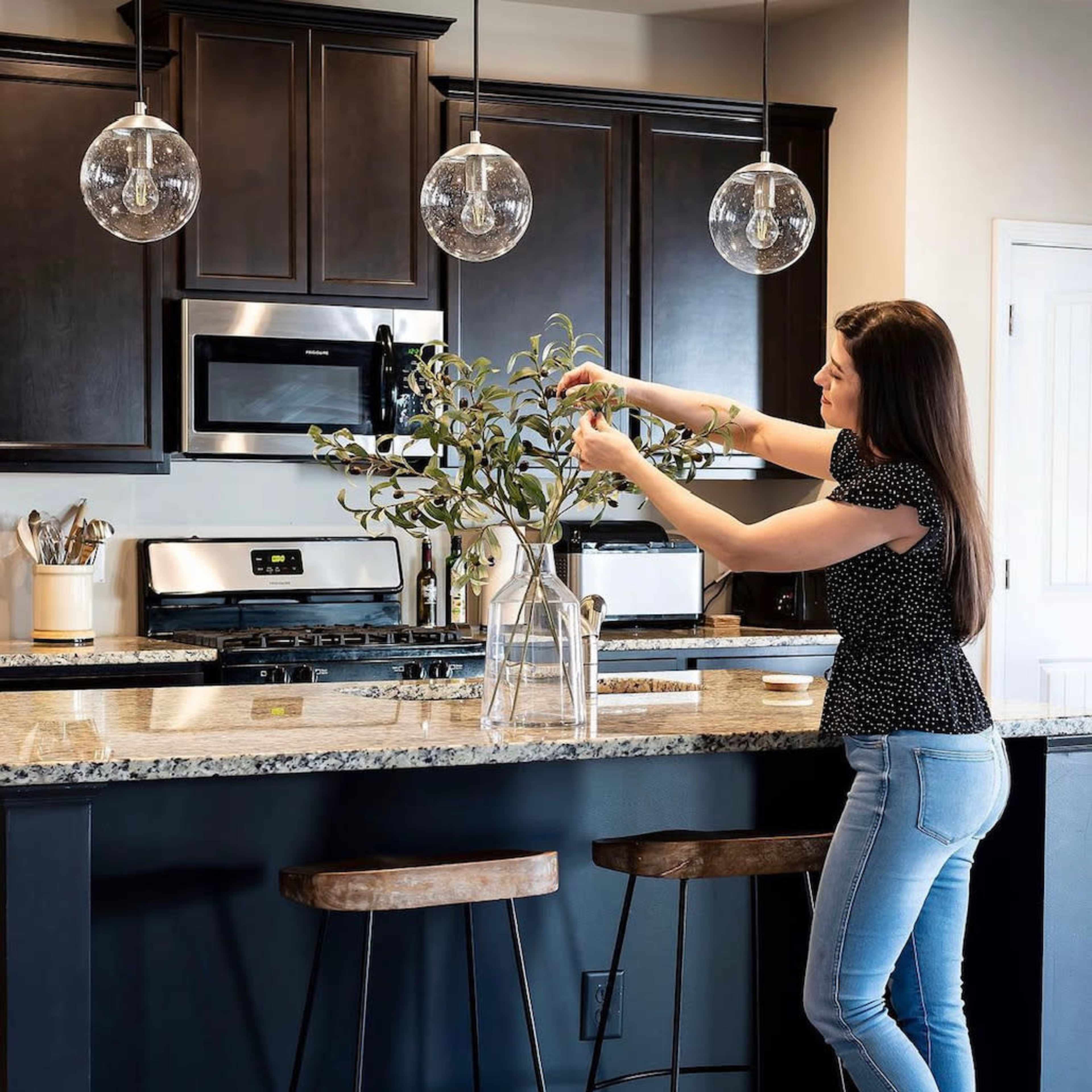 A woman arranges greenery in a glass vase on a kitchen island with dark wood cabinets and pendant lighting.