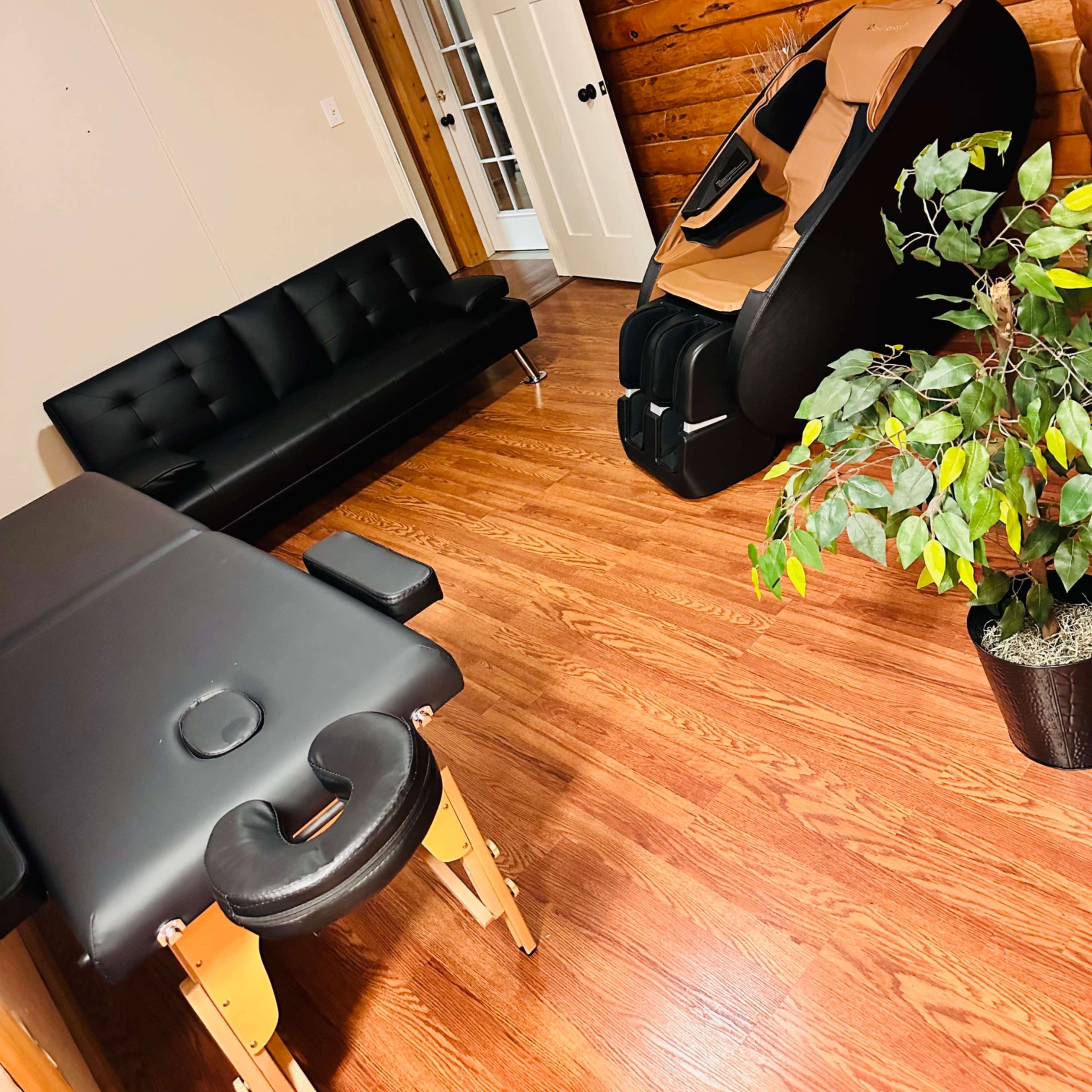 A room with wooden log walls, featuring a black sofa, a massage table, a massage chair, and a potted plant.