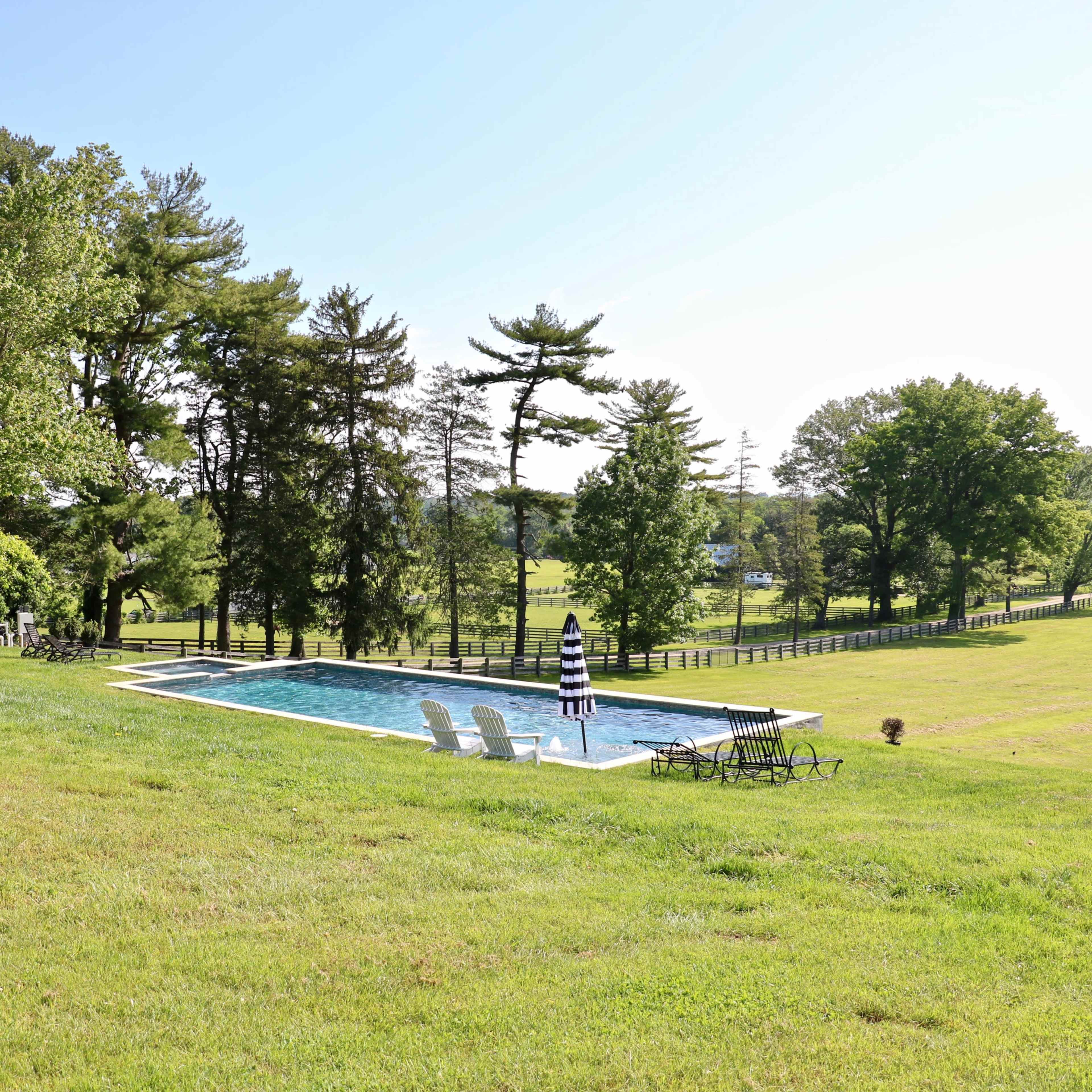 A swimming pool is situated on a grassy area surrounded by trees and a fenced pasture.