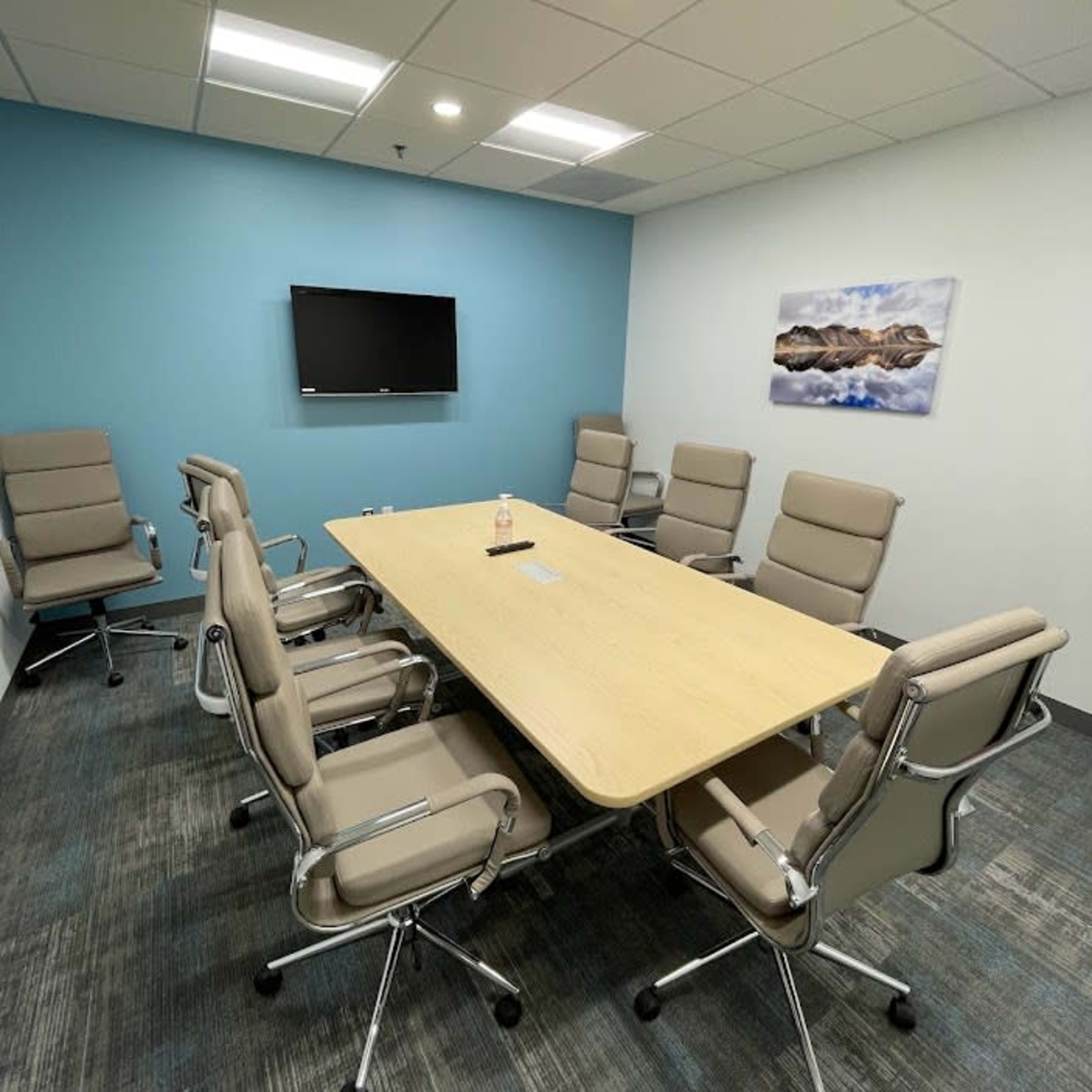 A conference room features a long wooden table surrounded by six beige chairs, with a television mounted on a blue wall and a landscape print hanging nearby.