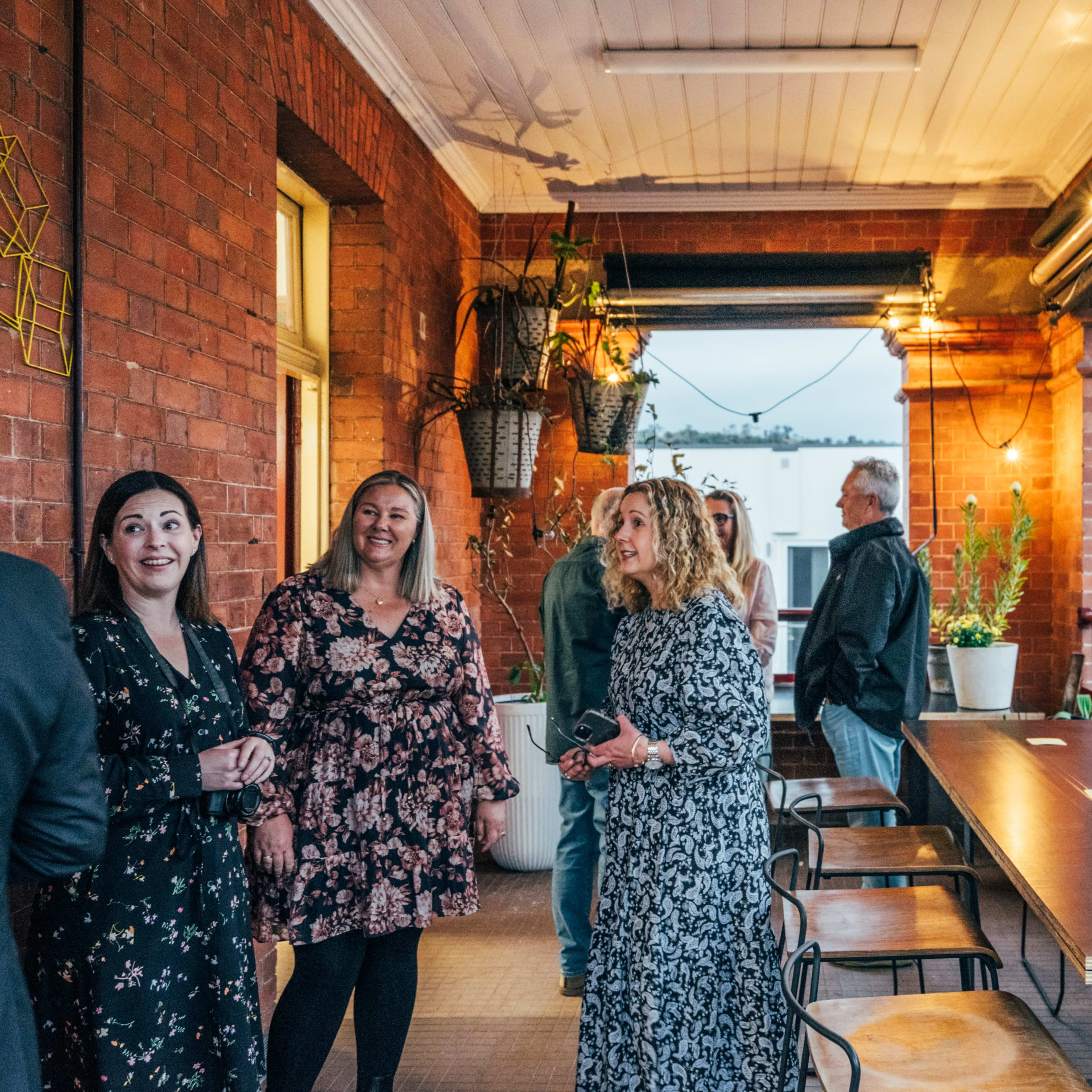 A group of people is standing and conversing on a balcony with brick walls and potted plants in the background.