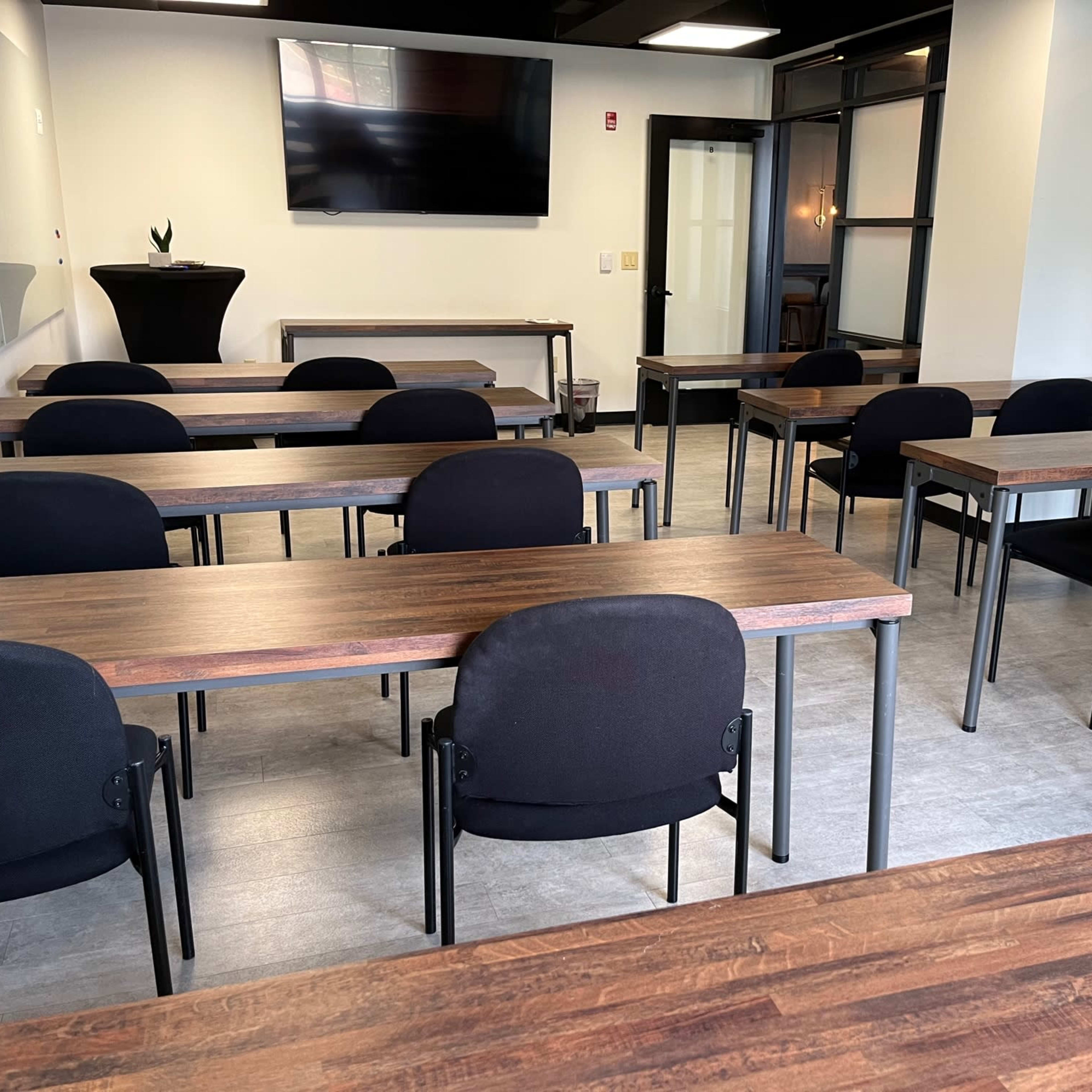 A classroom-style setup with several rows of wooden tables and black chairs arranged in a modern, well-lit space featuring a television screen on the wall.