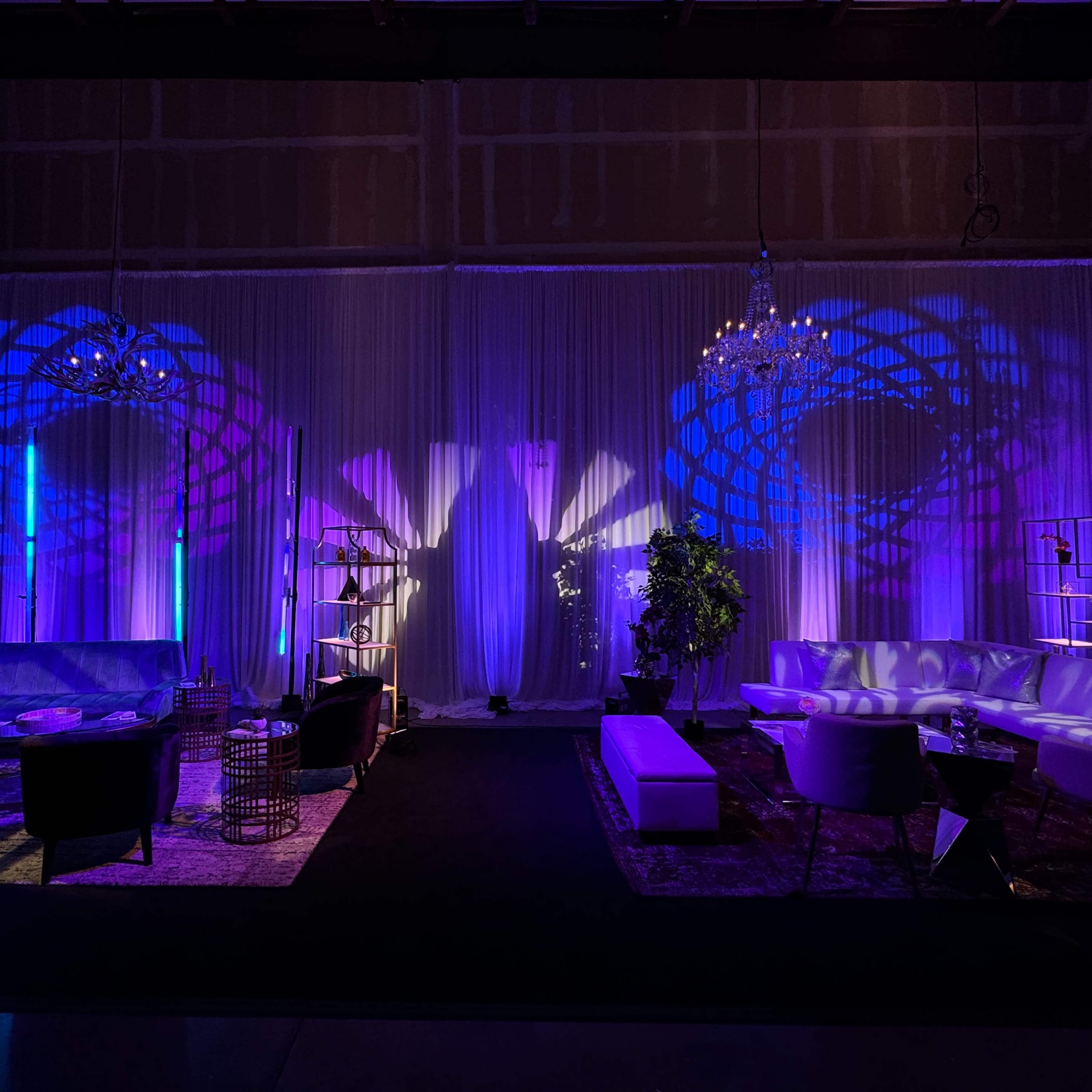 A stylish event space with a black carpet, white drapes, and purple lighting illuminating the walls, creating intricate shadow patterns behind modern seating arrangements.