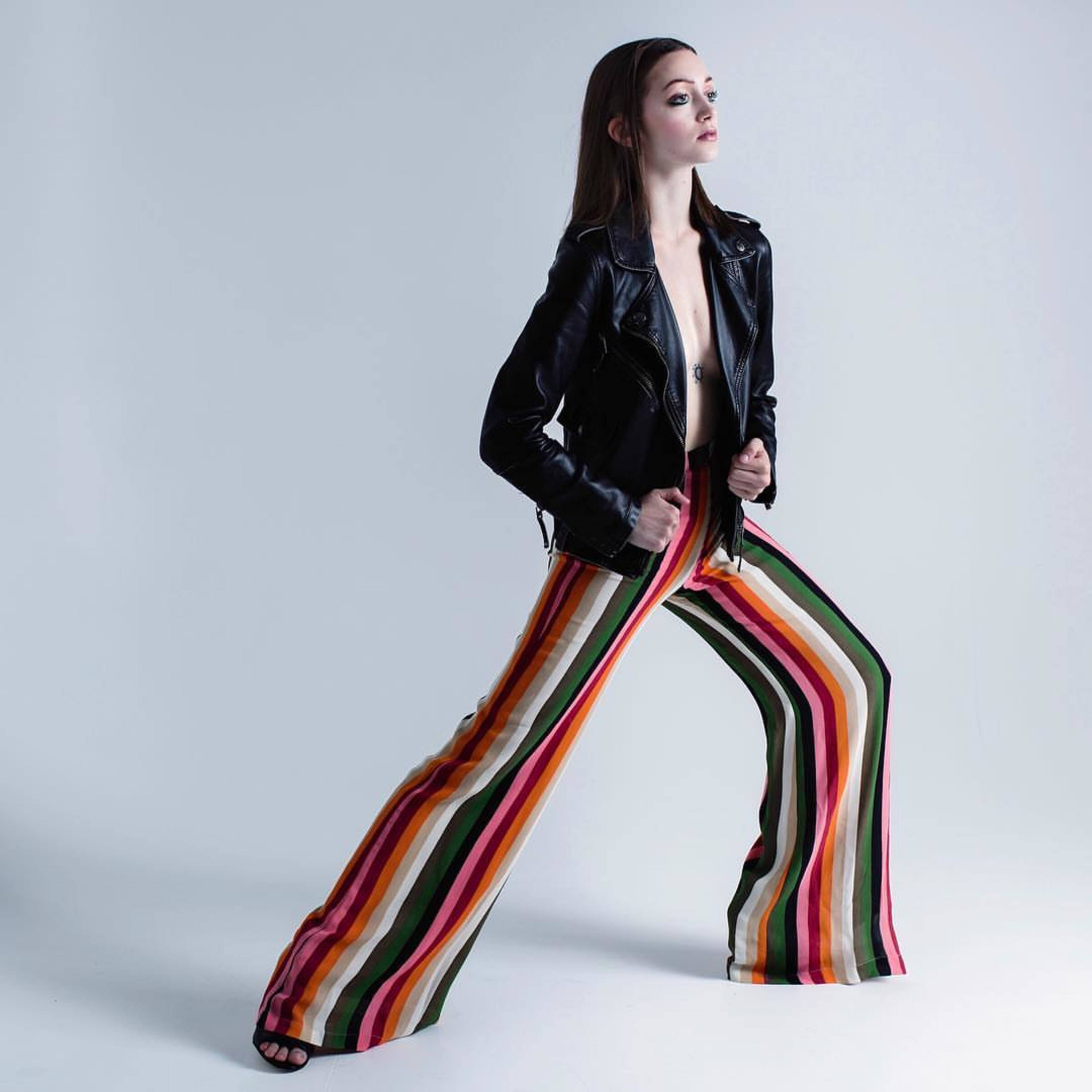 A model stands confidently wearing a black leather jacket and vibrant, striped wide-leg pants.