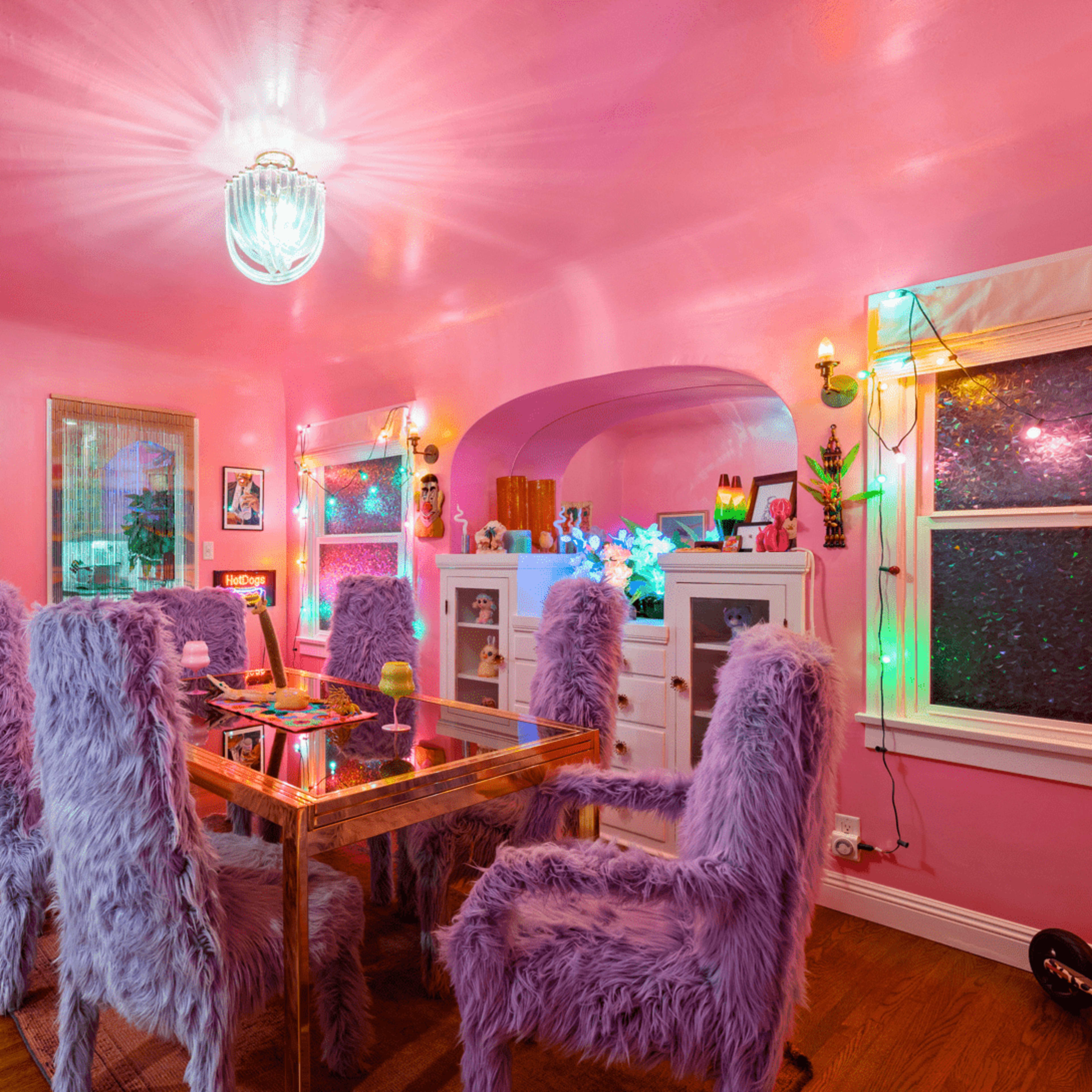 A dining room features a glass table surrounded by purple, fur-covered chairs, bright pink walls, and colorful decorations with a flamingo statue in one corner.
