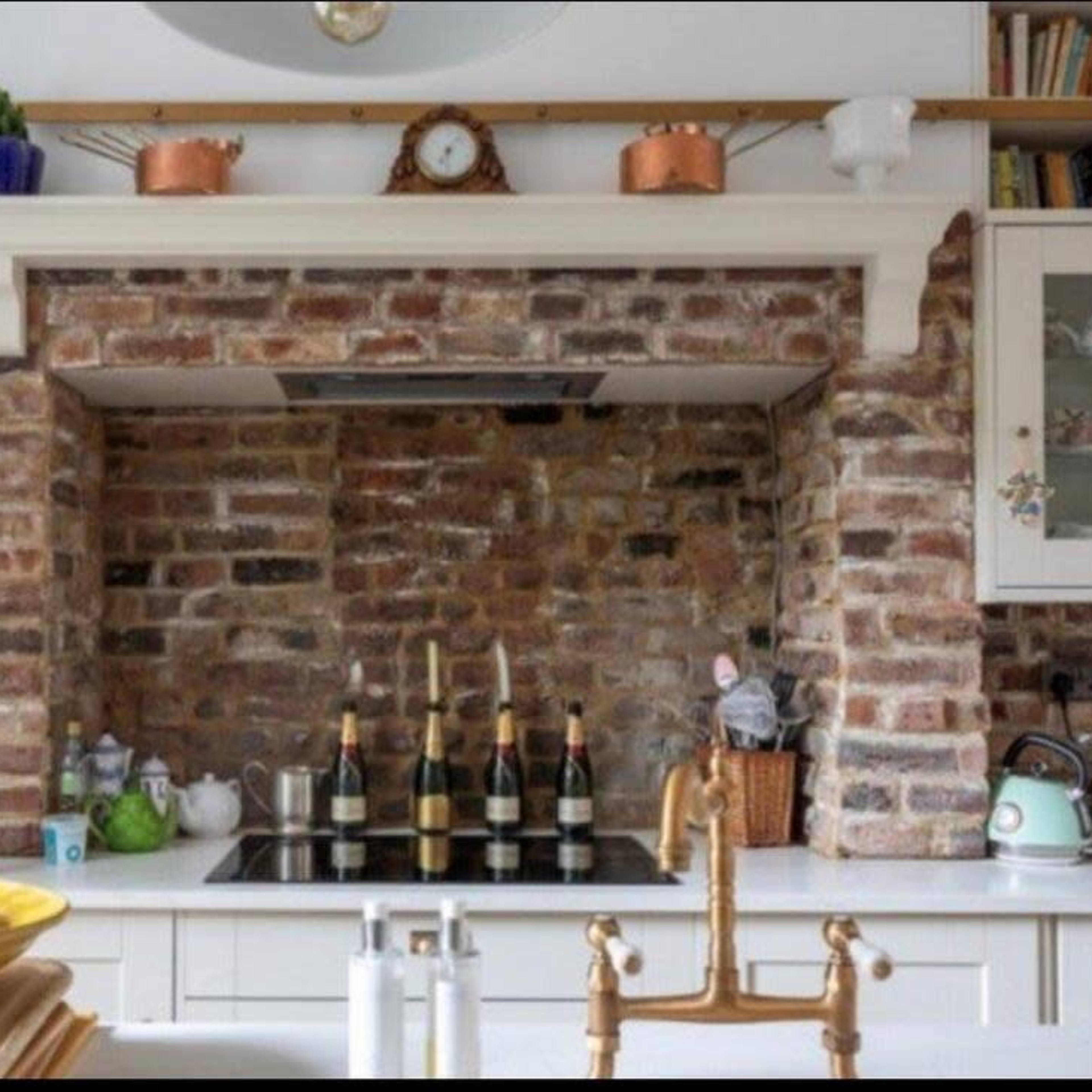 A rustic kitchen with a brick feature wall, a stove beneath a brick arch, and wooden shelves holding various kitchen items.