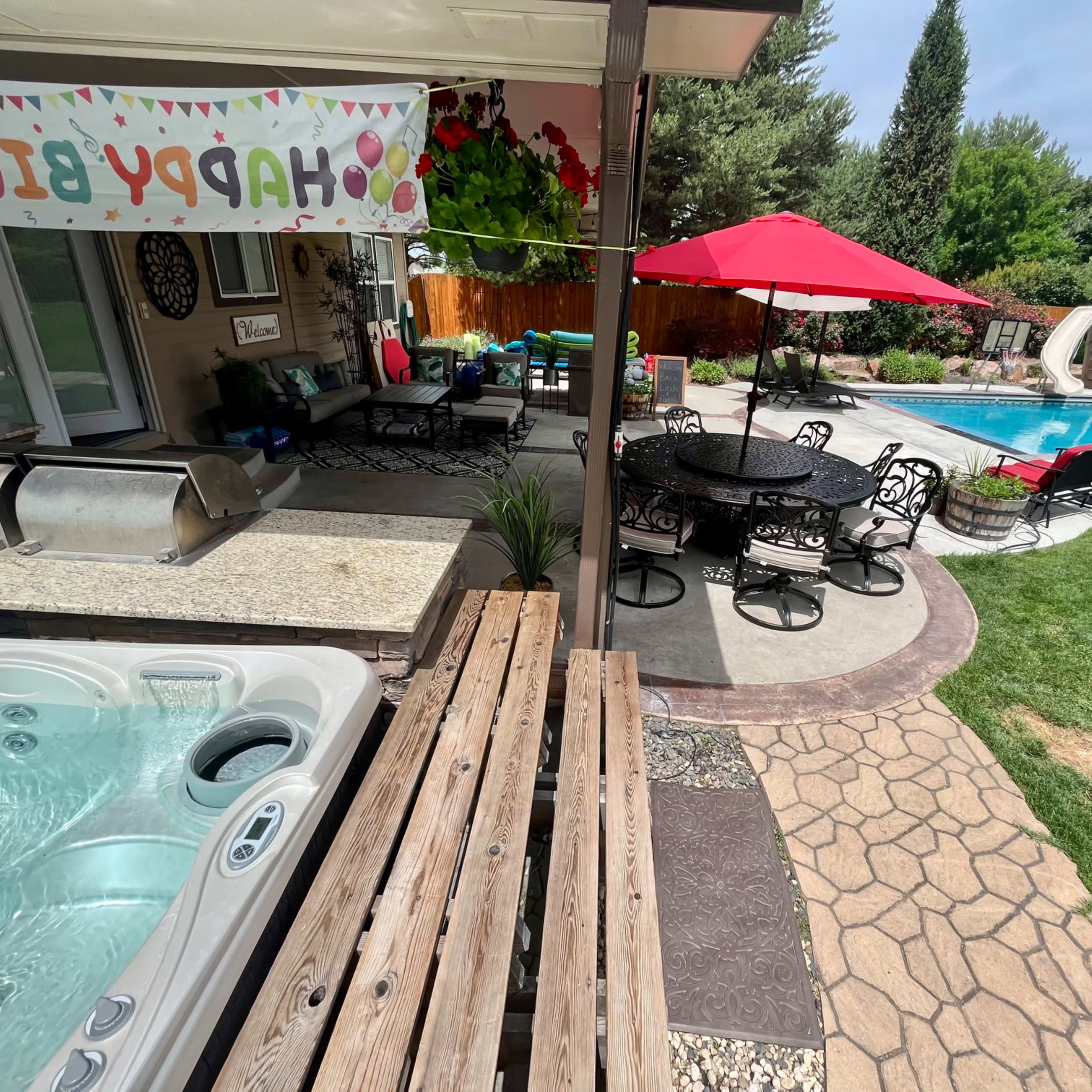 A backyard with a hot tub, a dining area under a canopy, a swimming pool, and a red umbrella beside green grass.