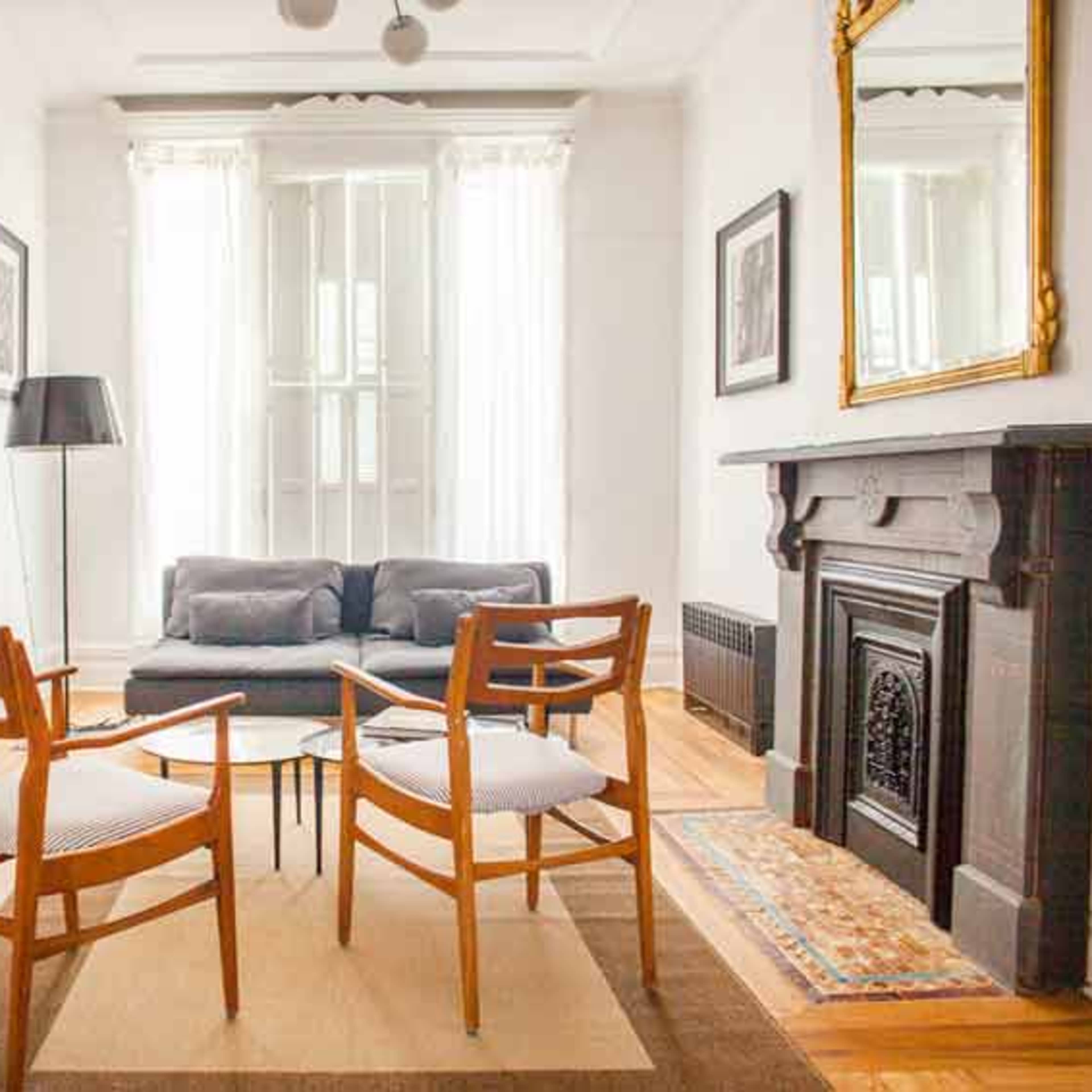 A bright living room featuring two wooden chairs facing a coffee table, a gray sofa, and a fireplace with a decorative mirror above it.