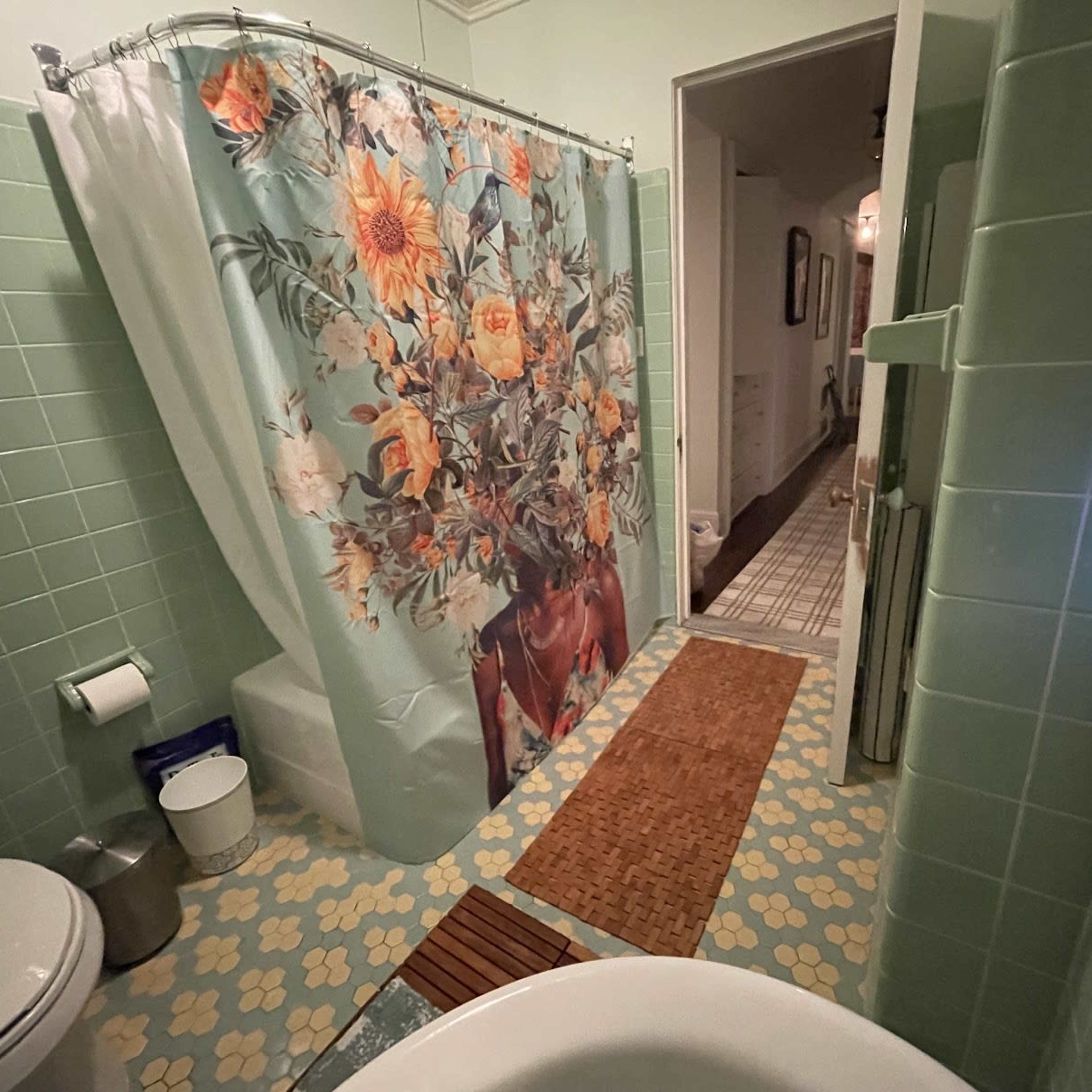 The bathroom features green tiled walls, a floral shower curtain, and a brown mat on the floor.