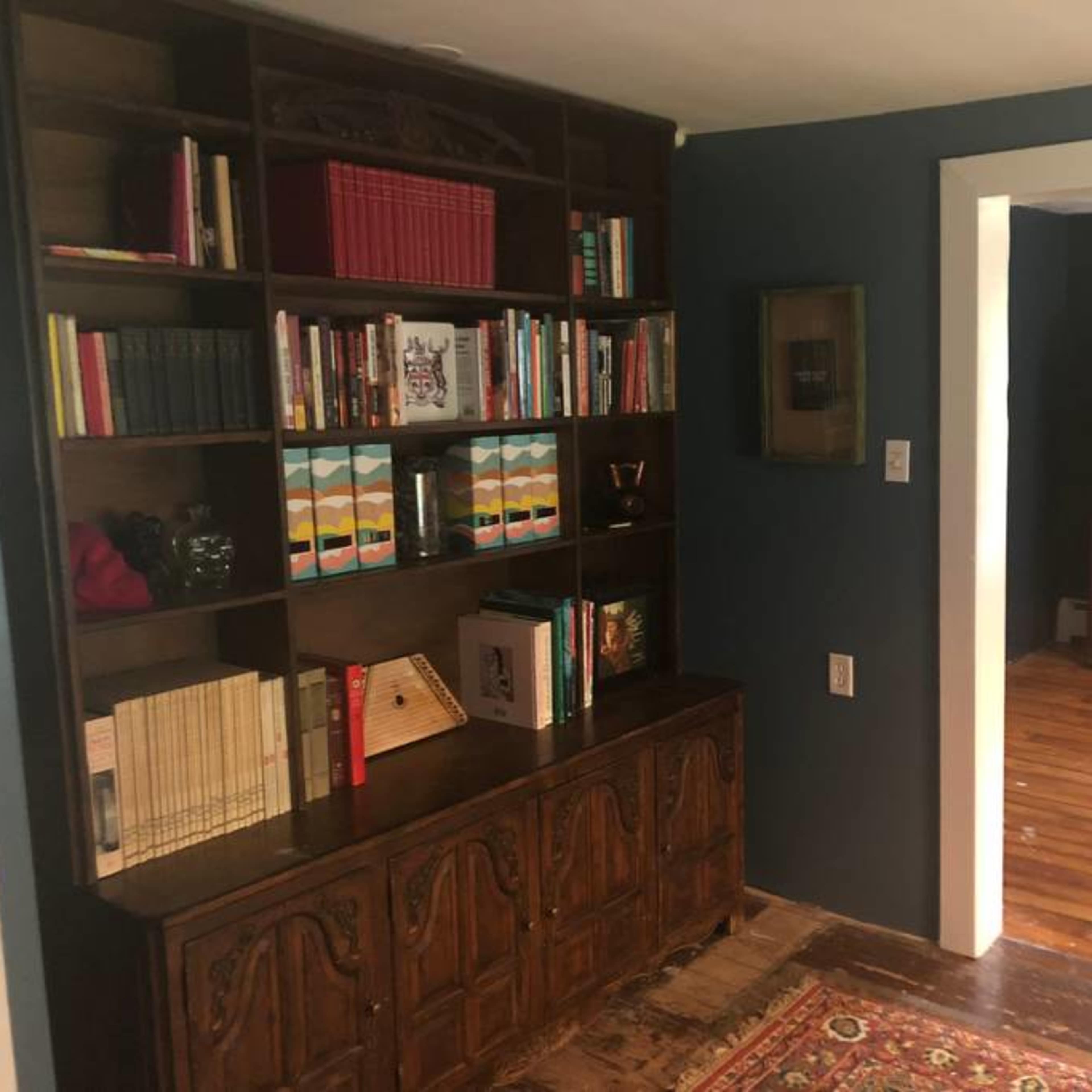 A wooden bookshelf filled with books and decorative items stands against a dark blue wall, with an open doorway leading to another room featuring a wooden wardrobe and exposed floorboards.
