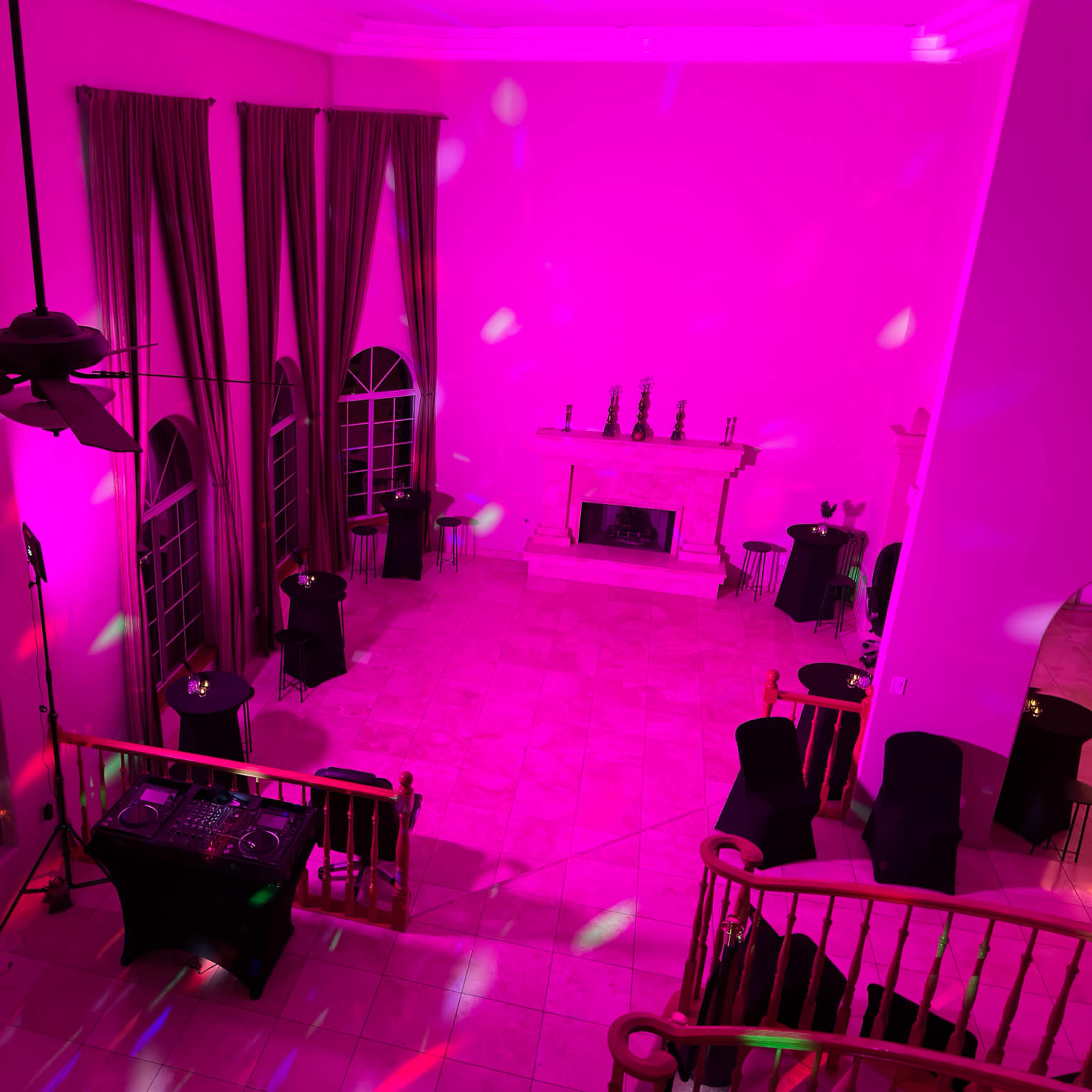 A brightly lit event space with pink lighting, featuring black tables and chairs arranged around a central area, along with a fireplace and tall windows.
