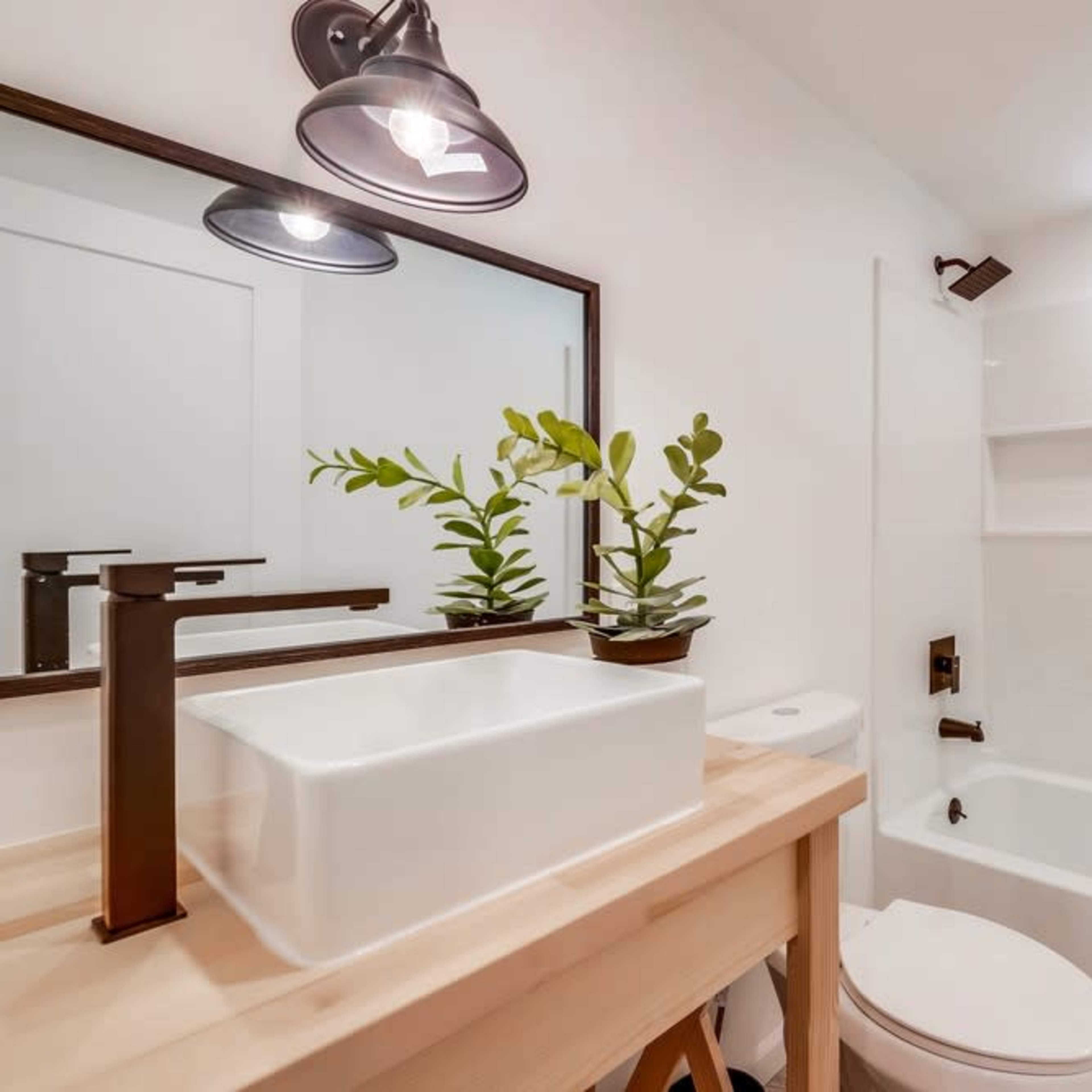 A clean, modern bathroom with a white bathtub, a sink on a wooden vanity, and a decorative plant on the counter.