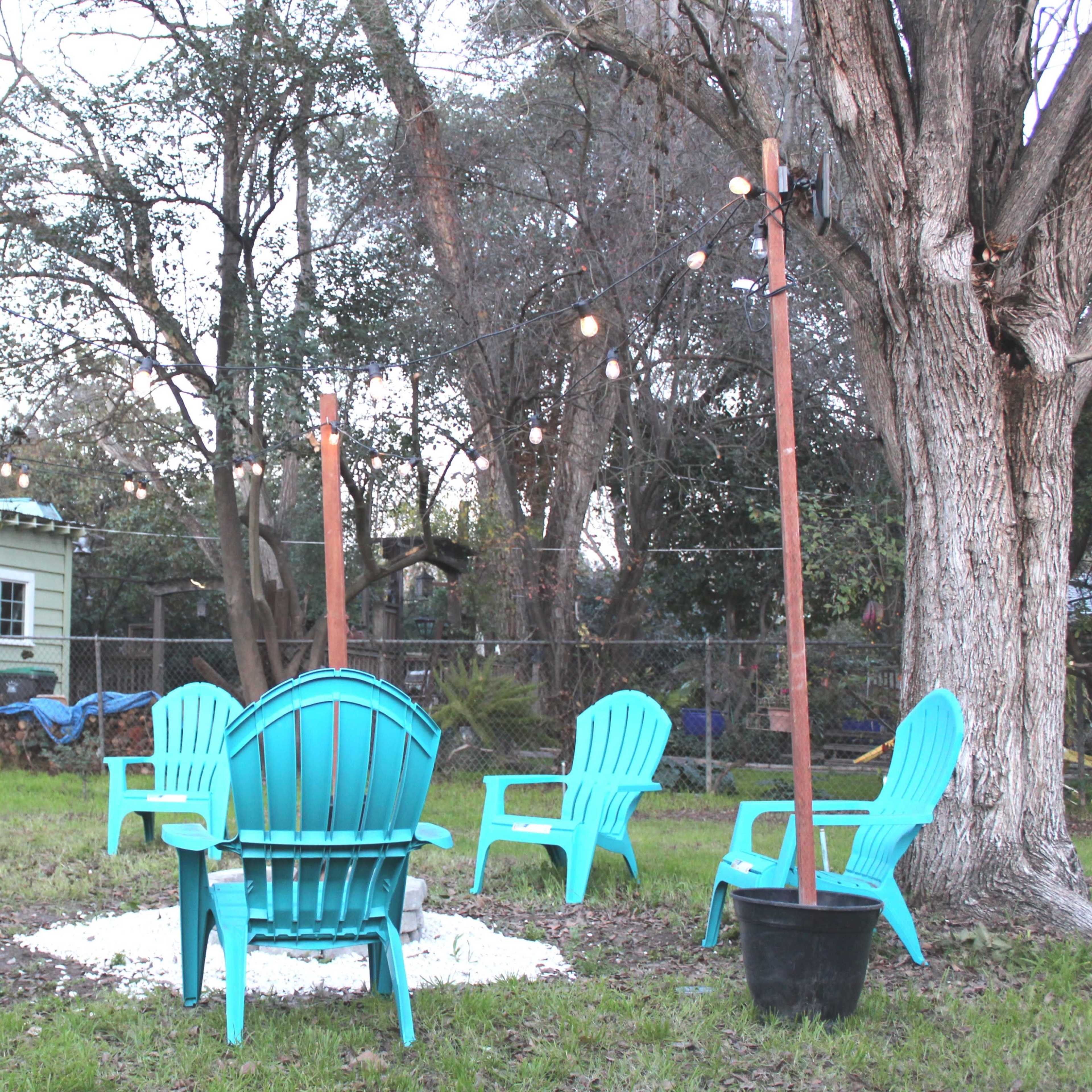A circular arrangement of turquoise Adirondack chairs surrounds a fire pit in a backyard, with string lights overhead and a house visible in the background.