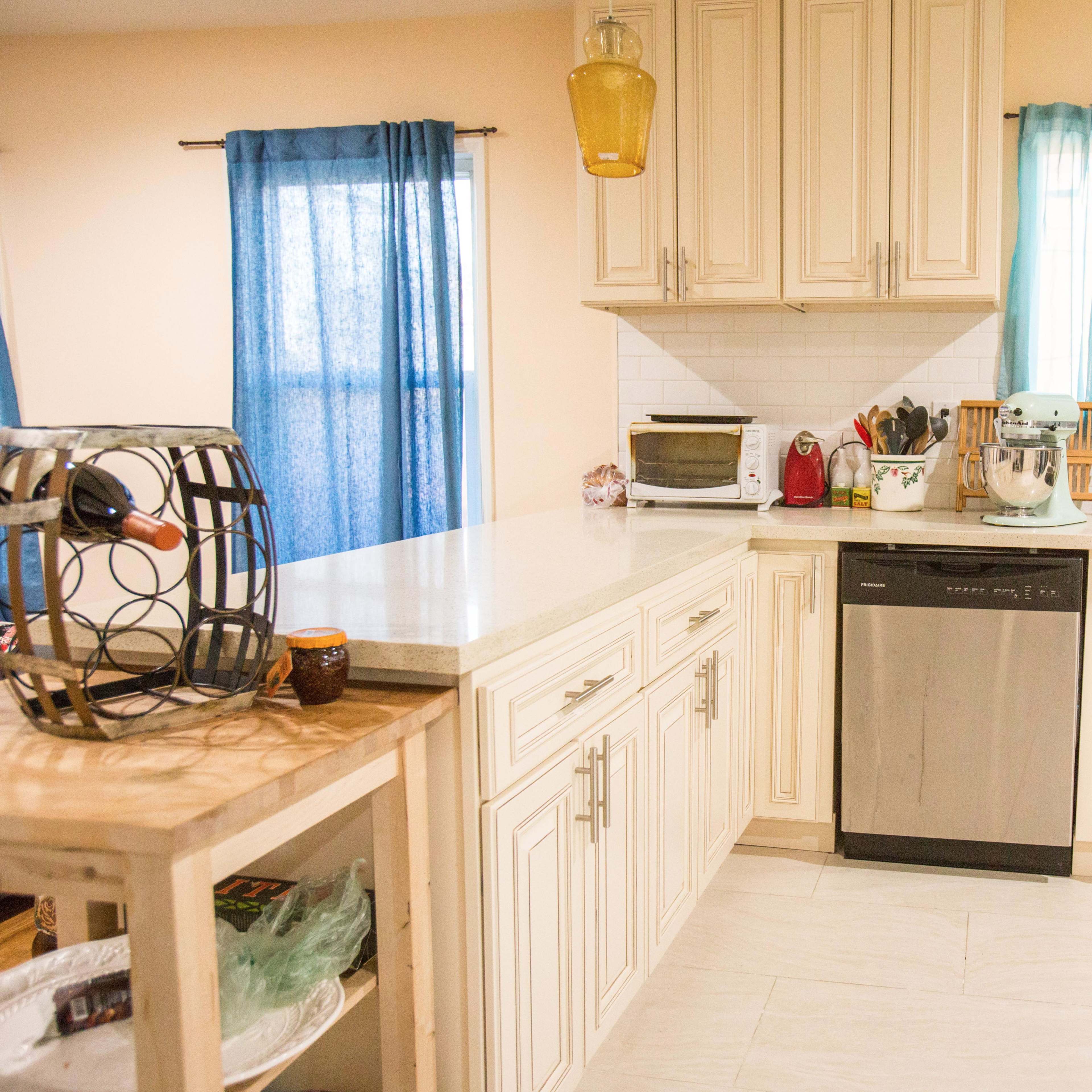 A modern kitchen features light-colored cabinets, stainless steel appliances, and blue curtains on the windows.