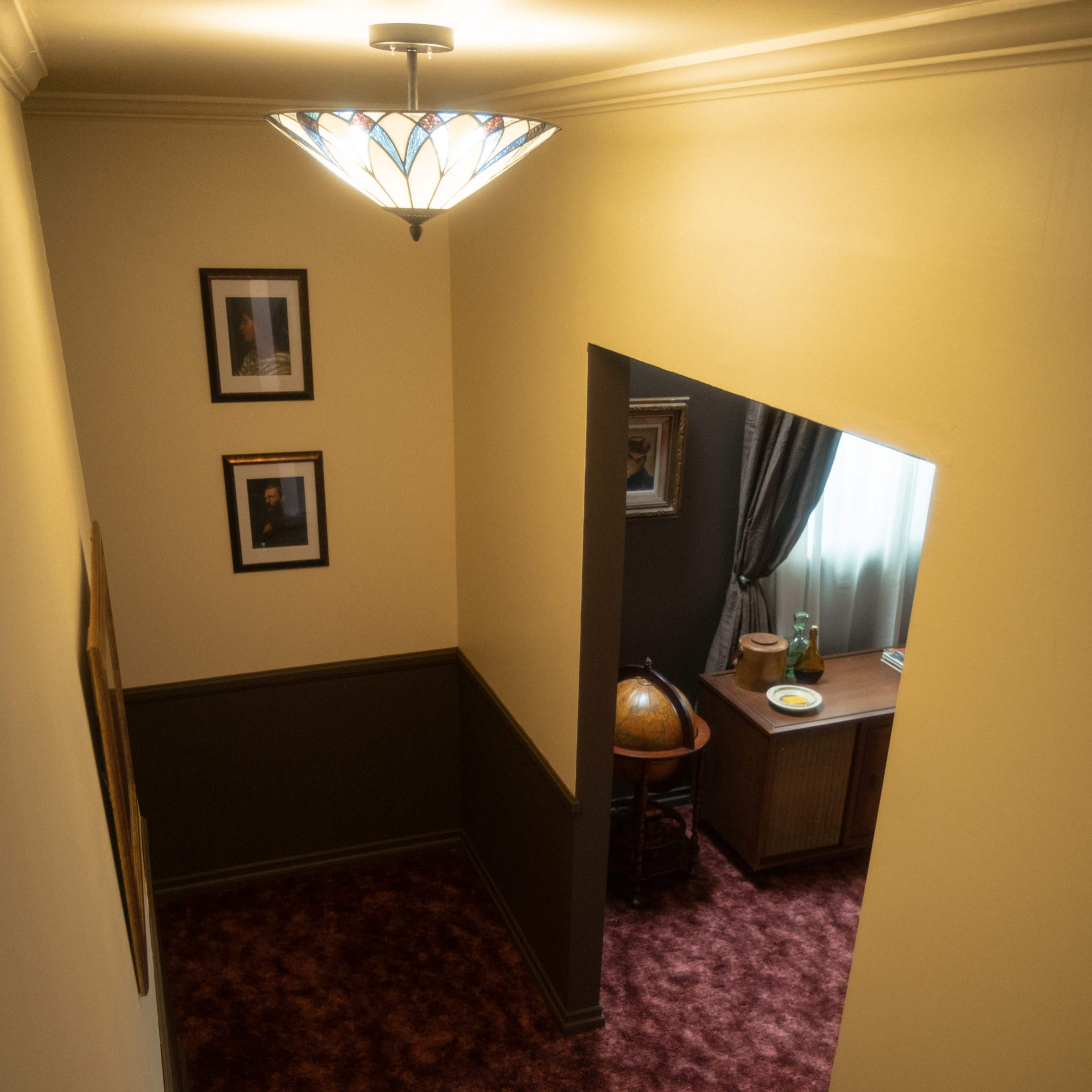 A hallway with a patterned carpet, framed pictures on the wall, and a small desk with a globe and decorative items in a room nearby.
