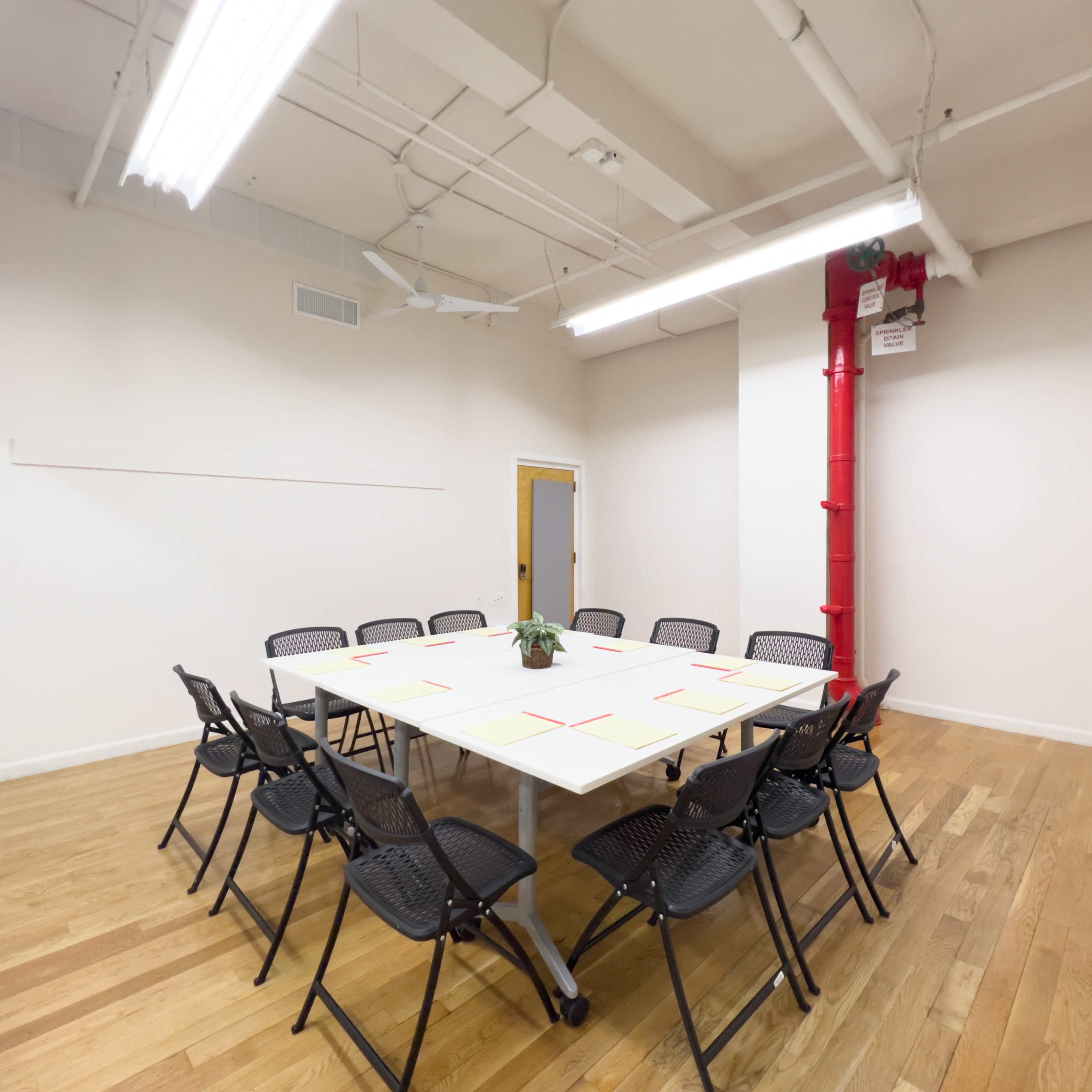 A rectangular table surrounded by black folding chairs sits in a minimalist meeting room with wooden flooring and a red pipe.