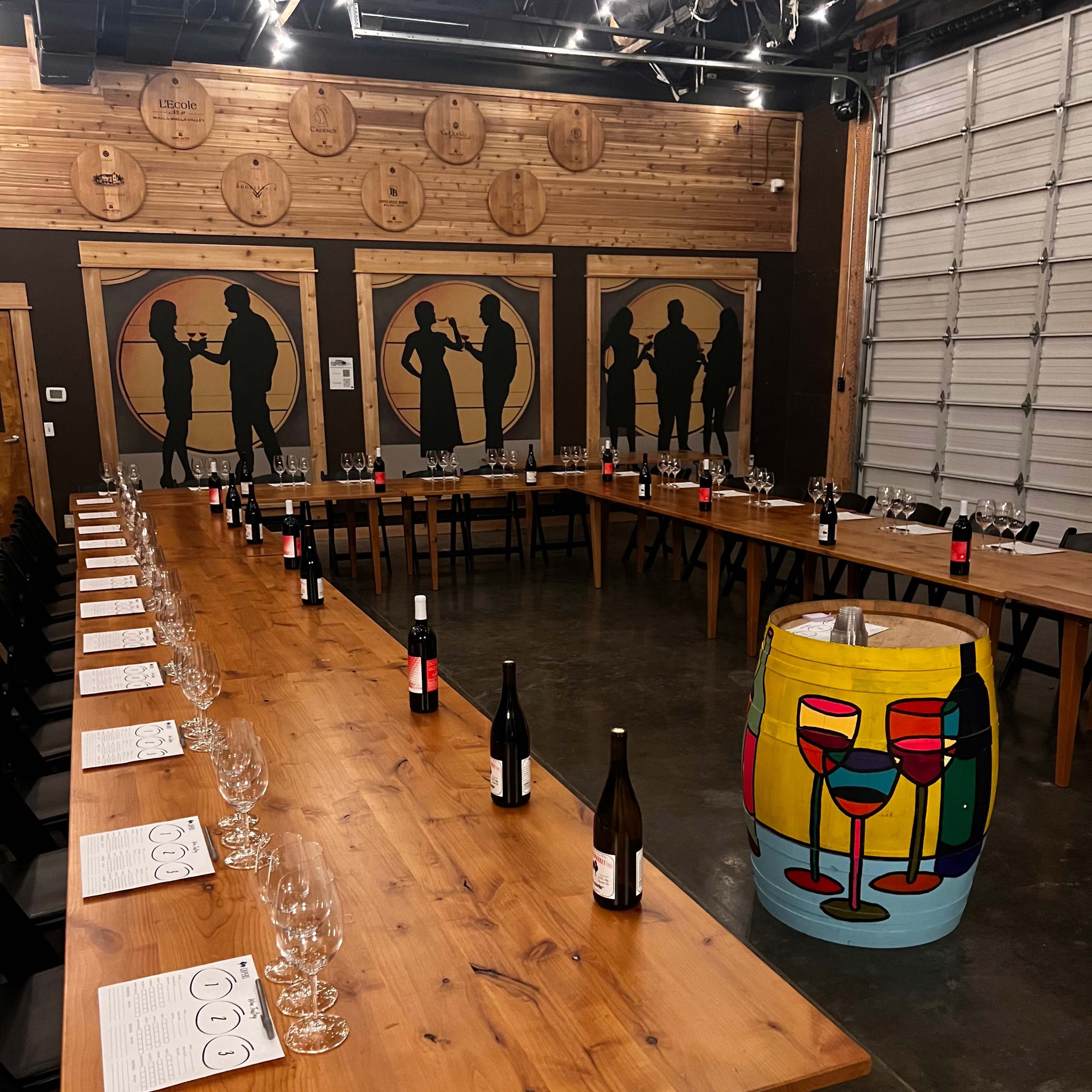 A large wooden dinner table is arranged in a U-shape with wine glasses and bottles, surrounded by silhouetted figures on the walls and a colorful painted barrel at one end.