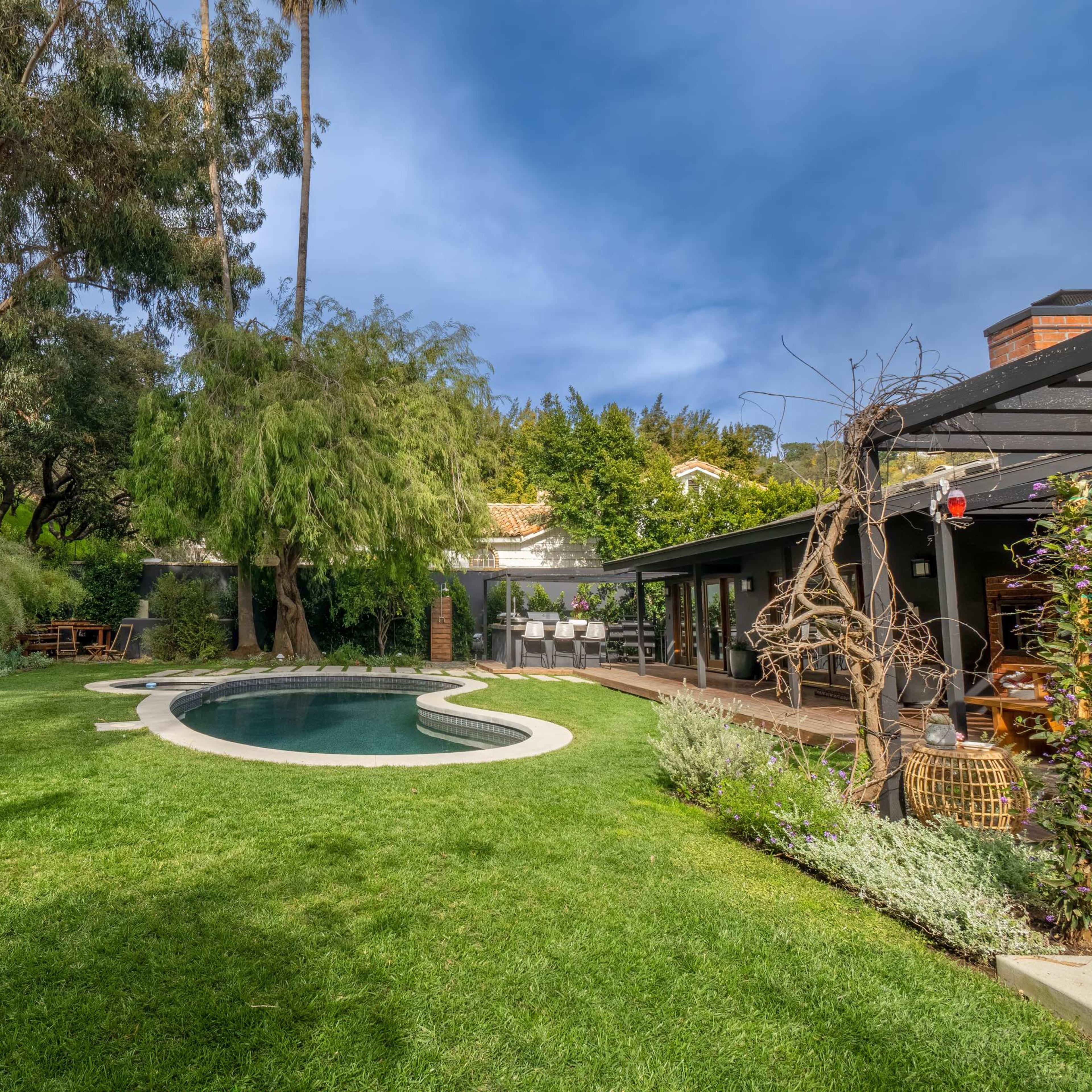 A landscaped backyard features a swimming pool surrounded by grass and trees, with a wooden deck and seating area nearby.