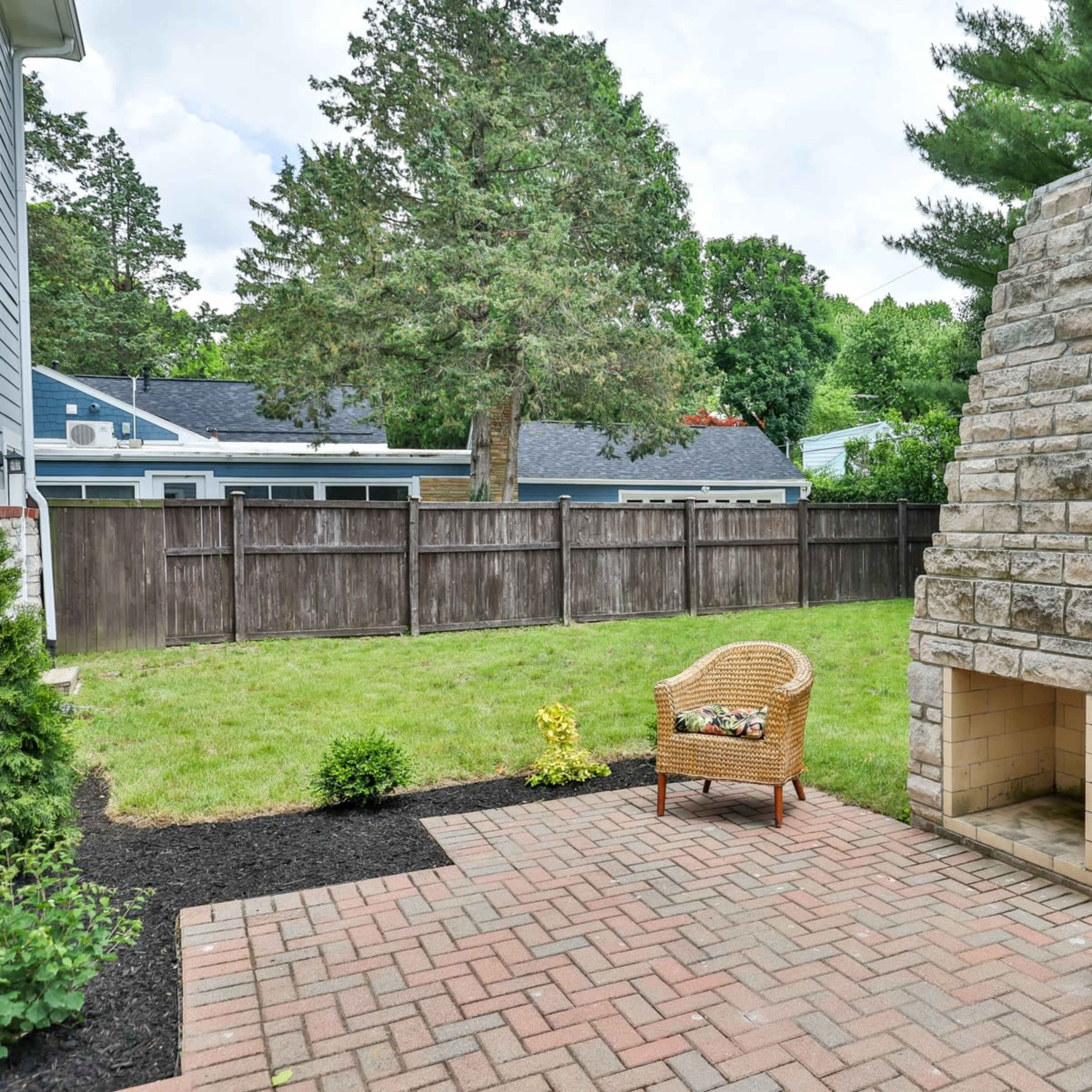 A backyard patio with two wicker chairs facing a stone outdoor fireplace and surrounded by a wooden fence and greenery.