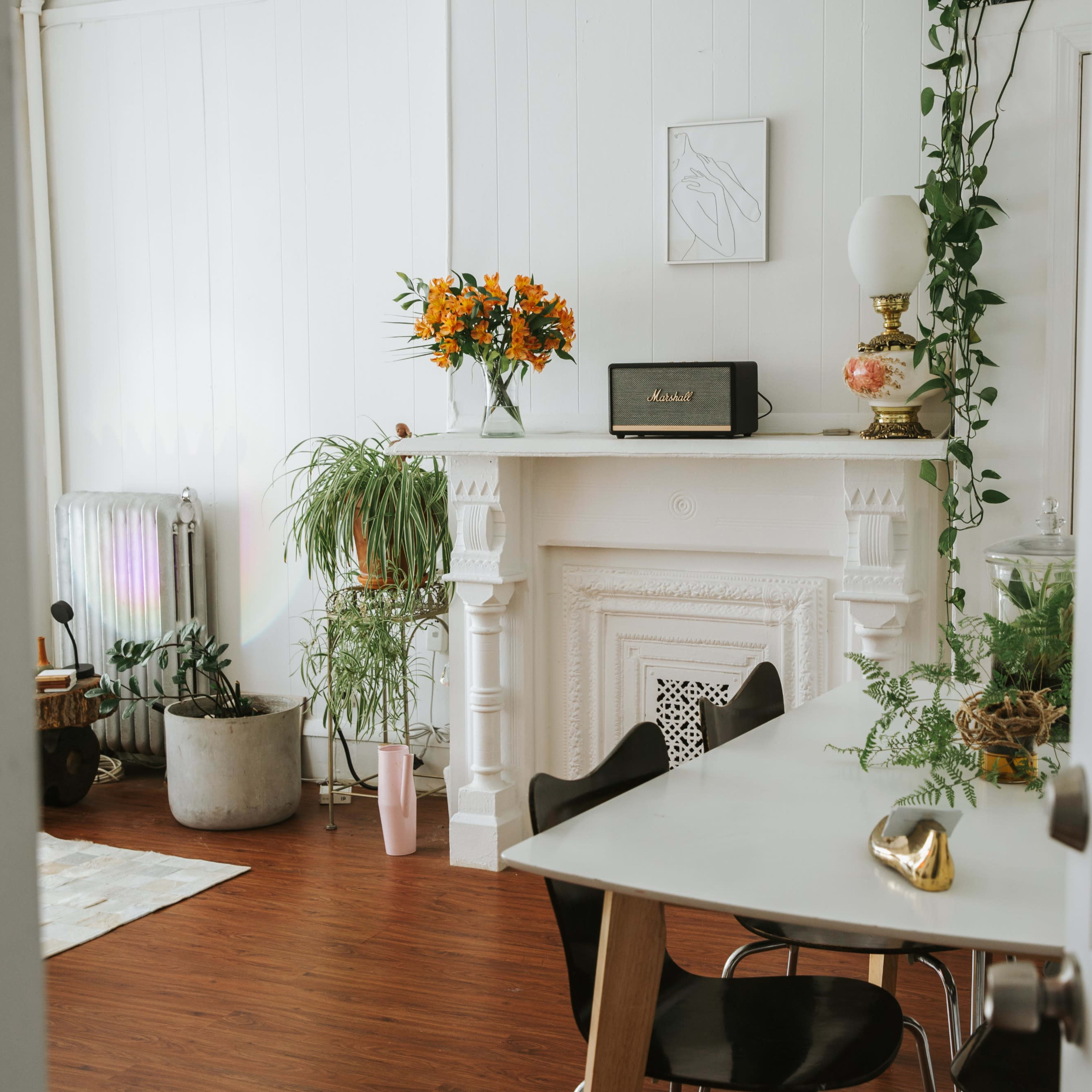 A bright room features a white fireplace adorned with flowers and plants, alongside a dining table and black chairs.