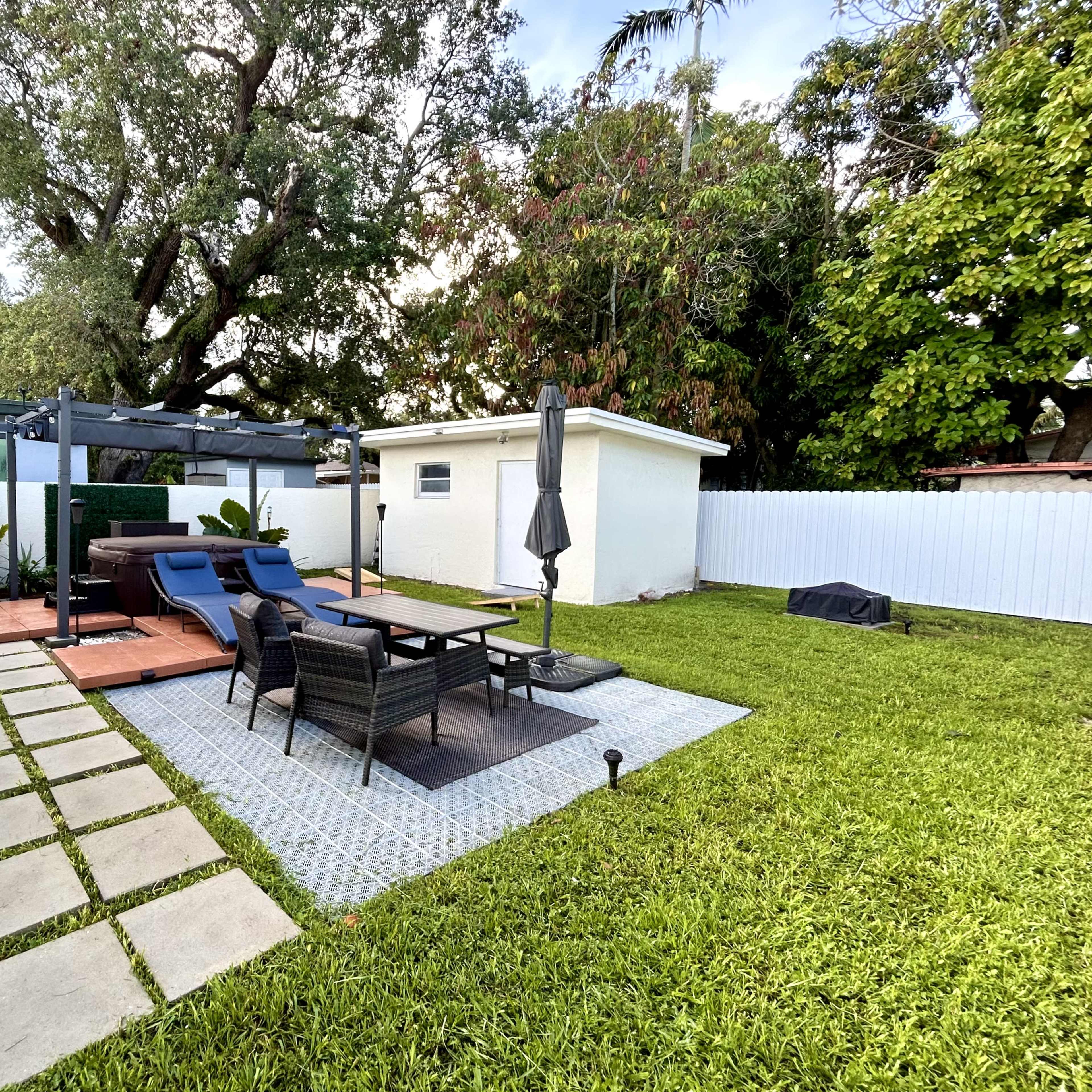 A backyard featuring a patio with lounge chairs, a table, and a small shed surrounded by greenery and a white fence.