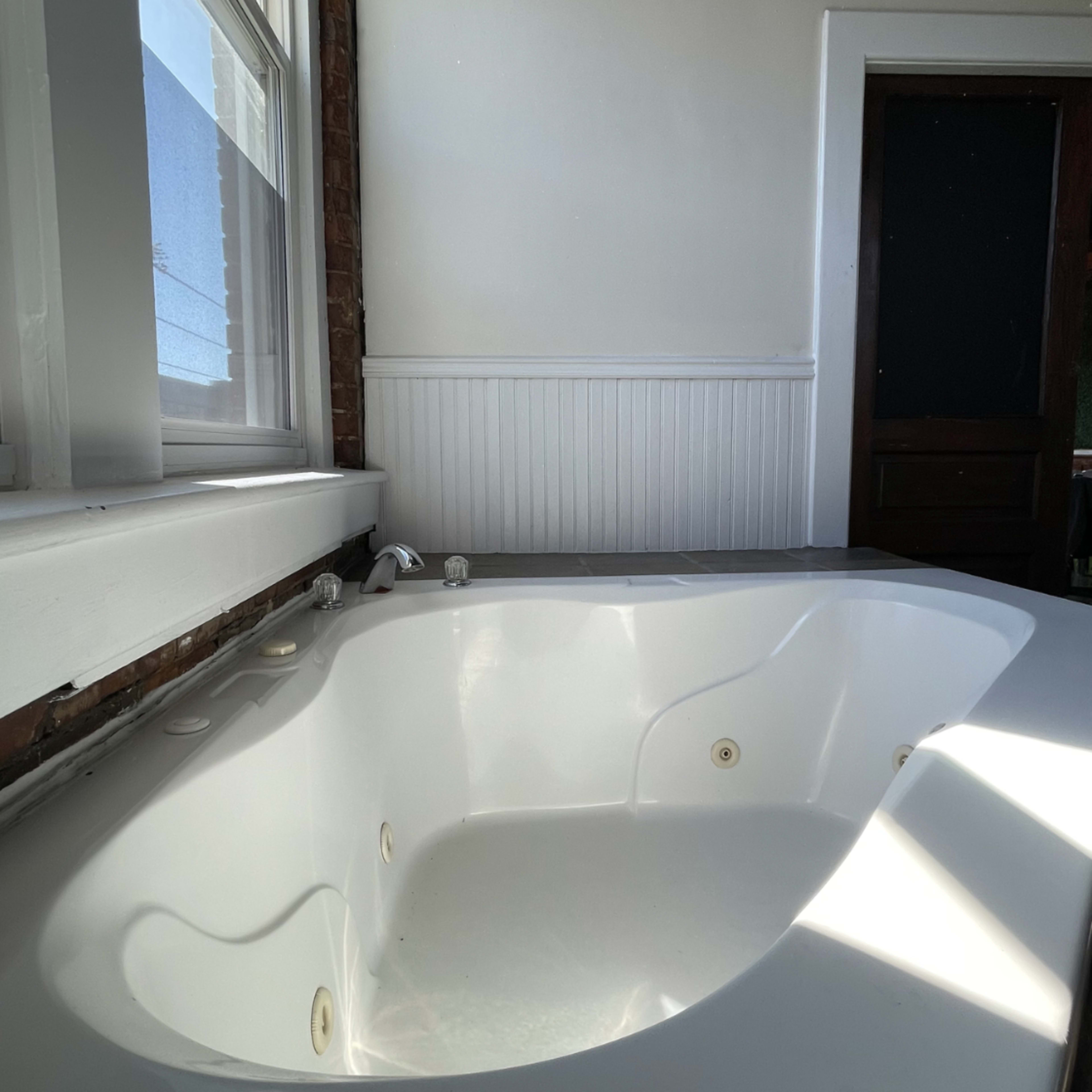 A white bathtub positioned near a window with a paneled wall and a dark wooden door in the background.