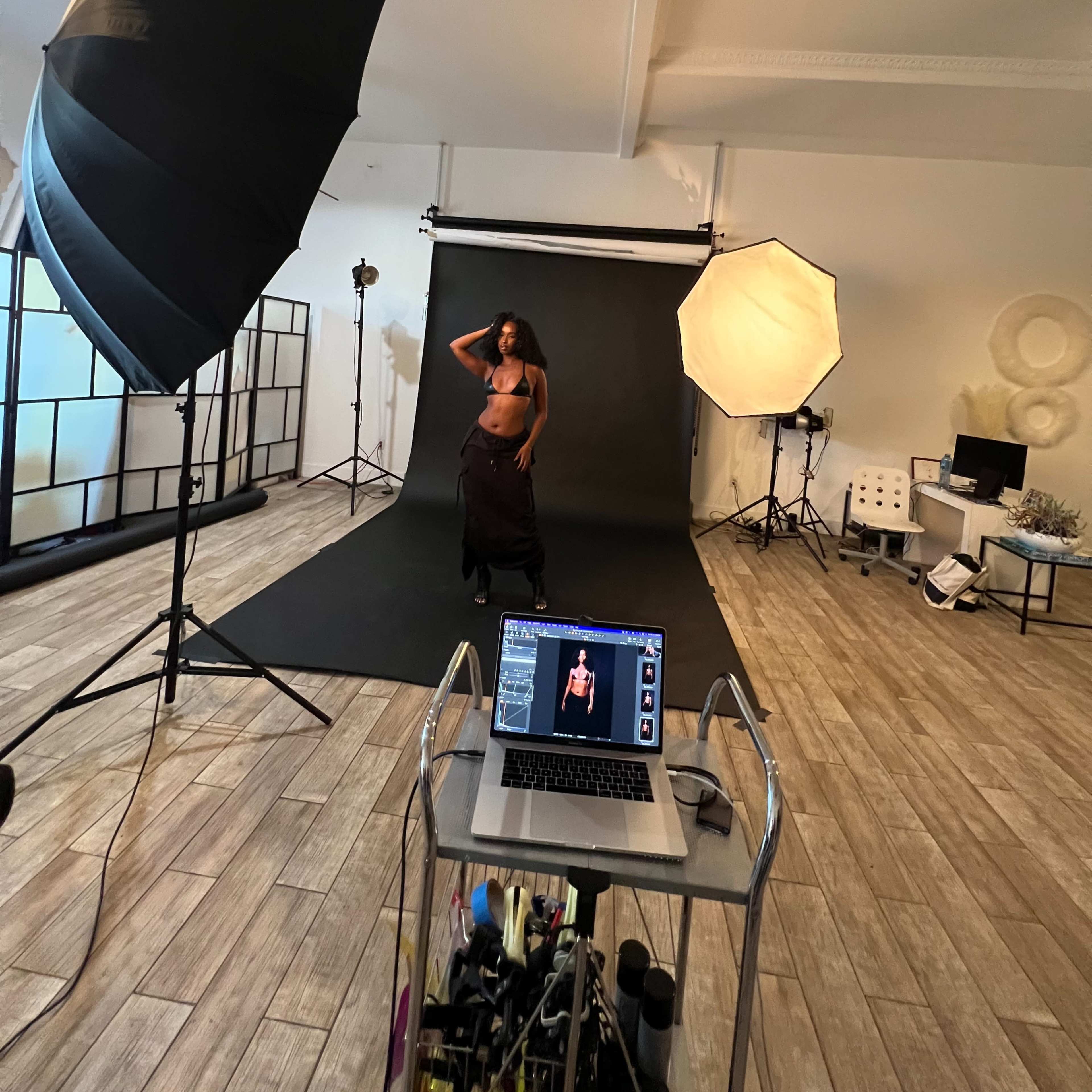 A model poses in front of a backdrop in a photography studio, with lighting equipment and a computer displaying the shot in progress.
